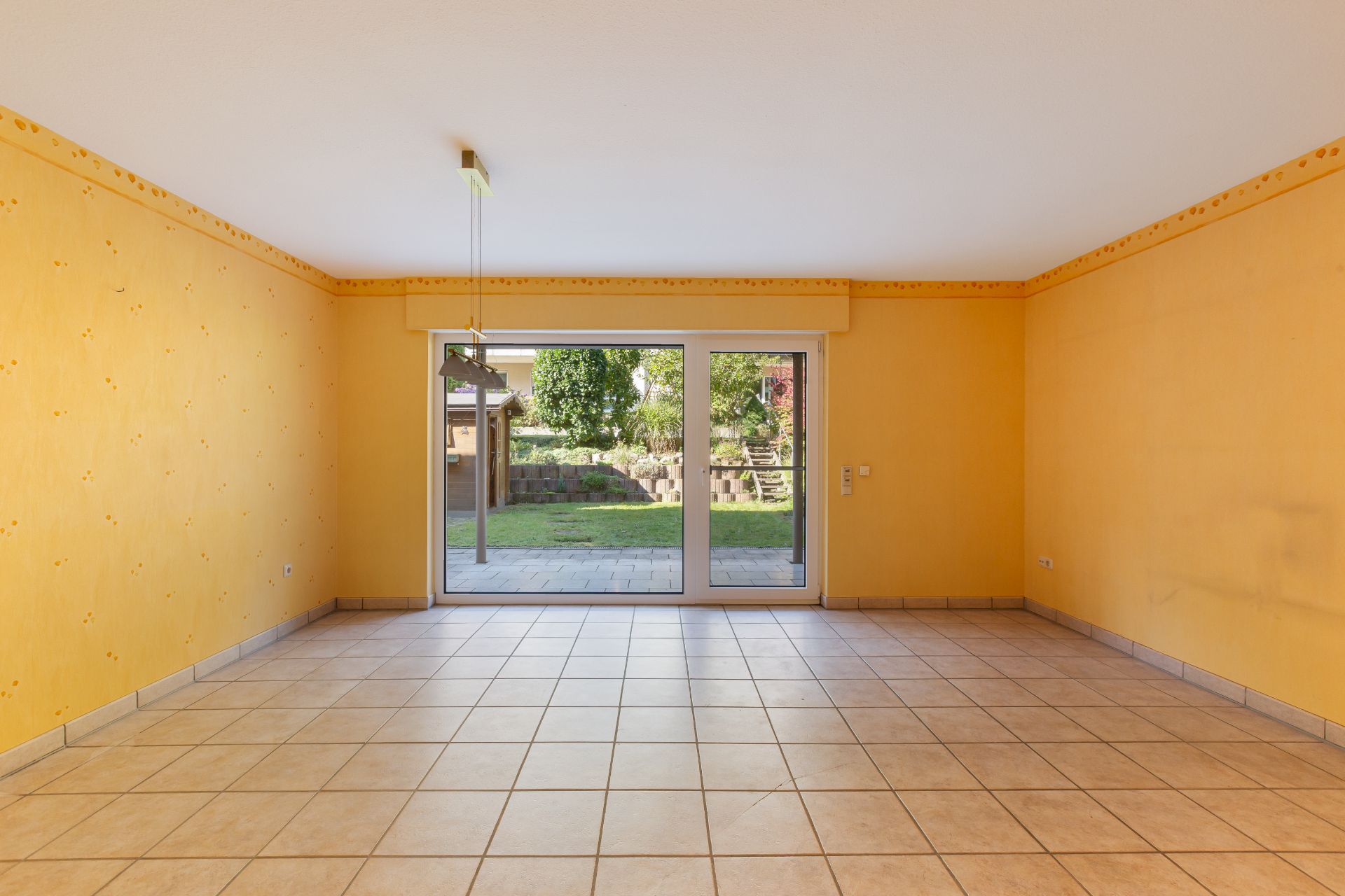Leerer Raum mit gelben Wänden, Fliesenboden und großer Glasschiebetür zum Garten.