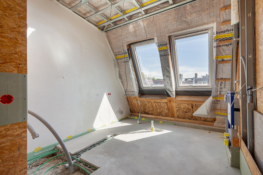 Zimmer im Dachgeschoss eines Wohnhauses im Rohbau mit zwei Dachfenstern und sichtbaren Leitungen.