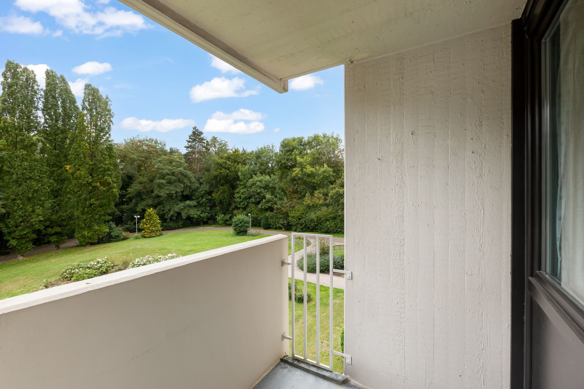 Balkon mit Blick auf einen grünen Garten und Bäume bei blauem Himmel.