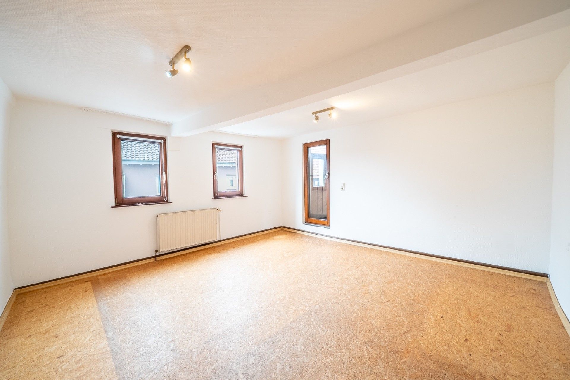 Leerer, heller Raum mit zwei Fenstern, einem Heizkörper und einer Glastür zu einem Balkon.