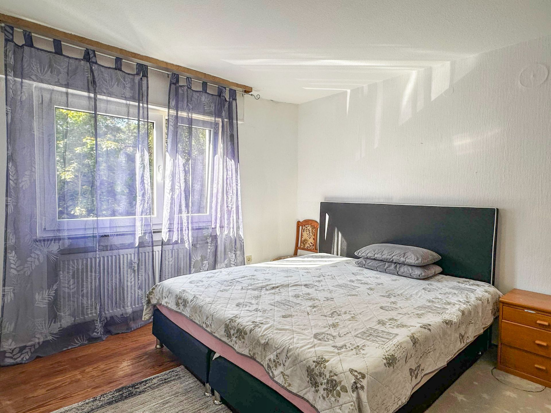 Helles Schlafzimmer mit Doppelbett, Nachttisch, Stuhl und Fenster mit transparentem Vorhang.