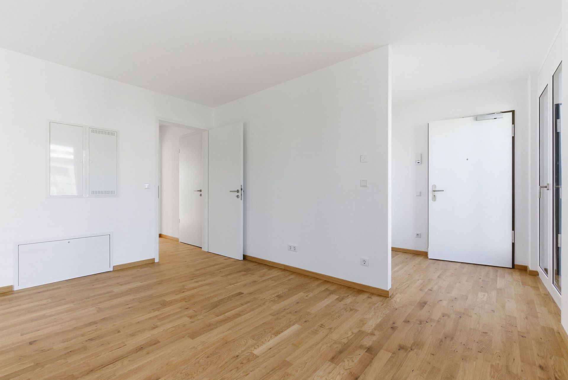 Helles, leeres Zimmer mit Holzboden, weißen Wänden und offenen Türen.