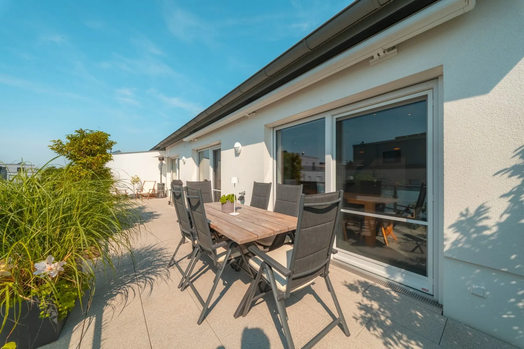Große Terrasse mit Holztisch, sechs Stühlen und Pflanzen neben einem Wohnhaus.