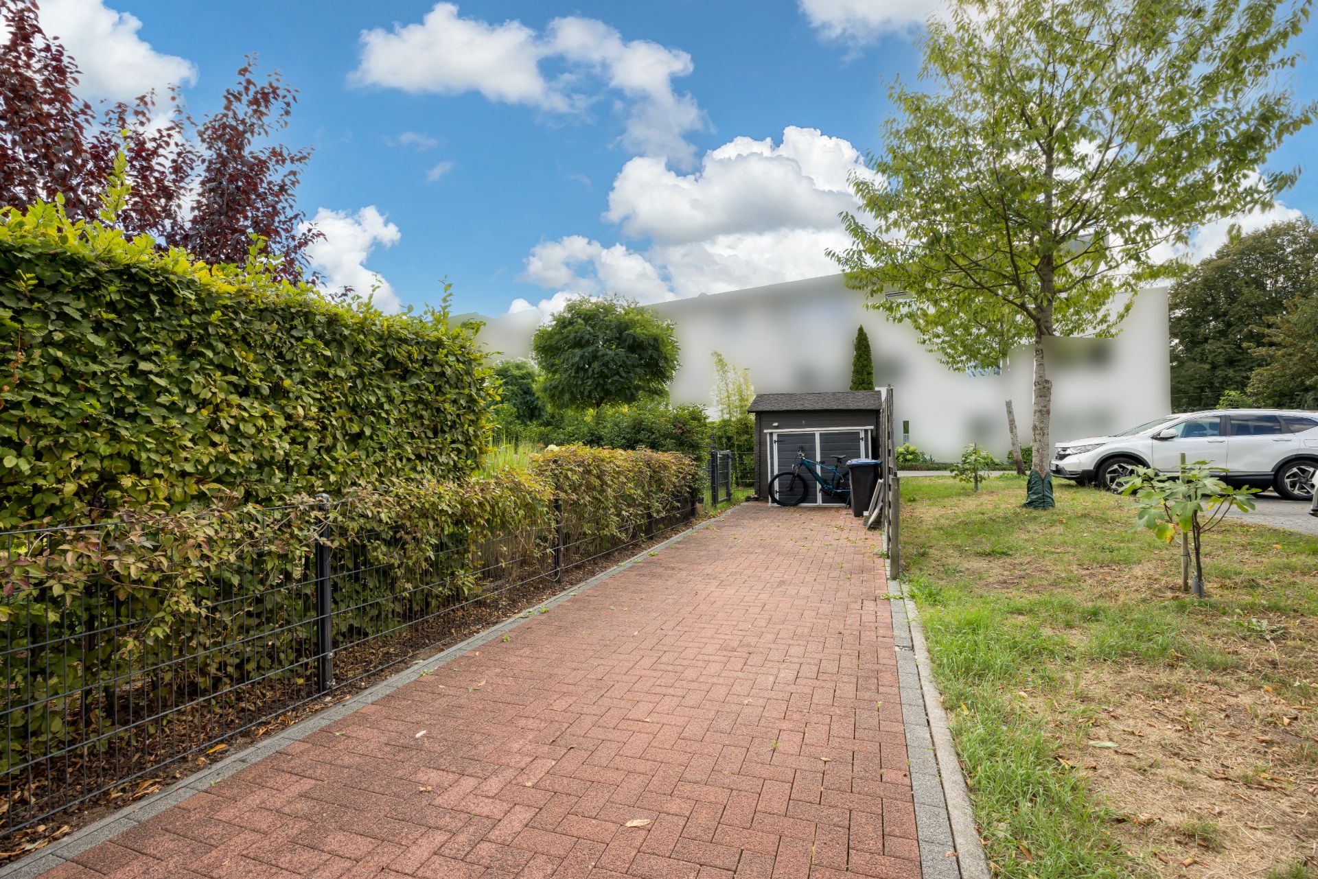 Roter gepflasterter Weg mit Hecke links, Rasen rechts und einem kleinen Schuppen am Ende.