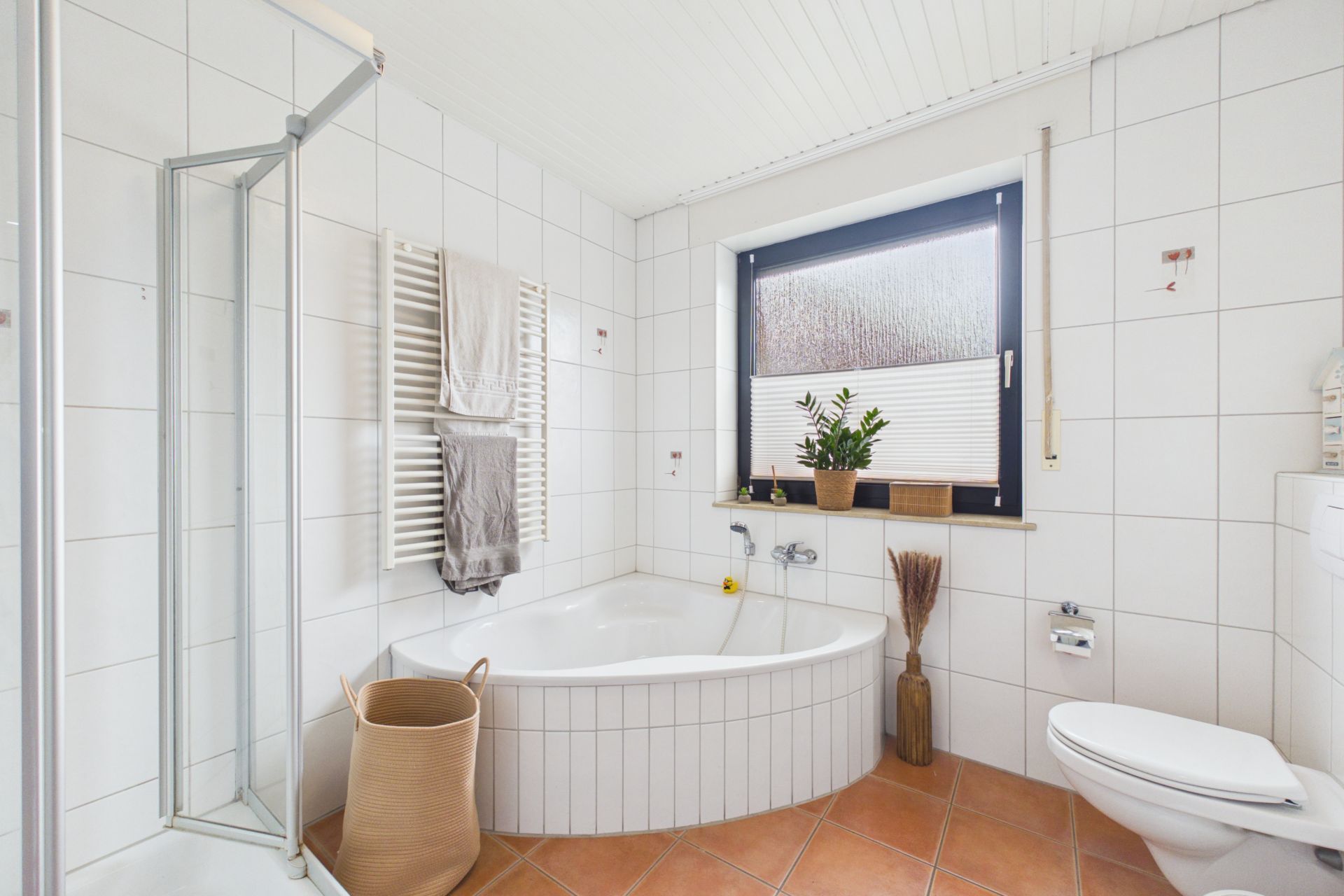 Modernes Badezimmer mit Eckbadewanne, Dusche, WC, Fenster und Pflanzen auf der Fensterbank.