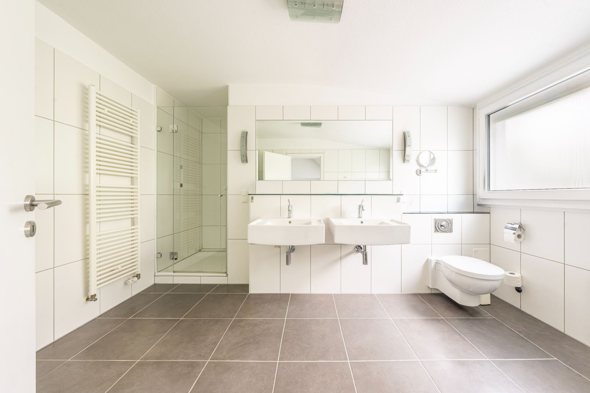 Modernes, weiß gefliestes Badezimmer mit zwei Waschbecken, Dusche, WC und großem Fenster.