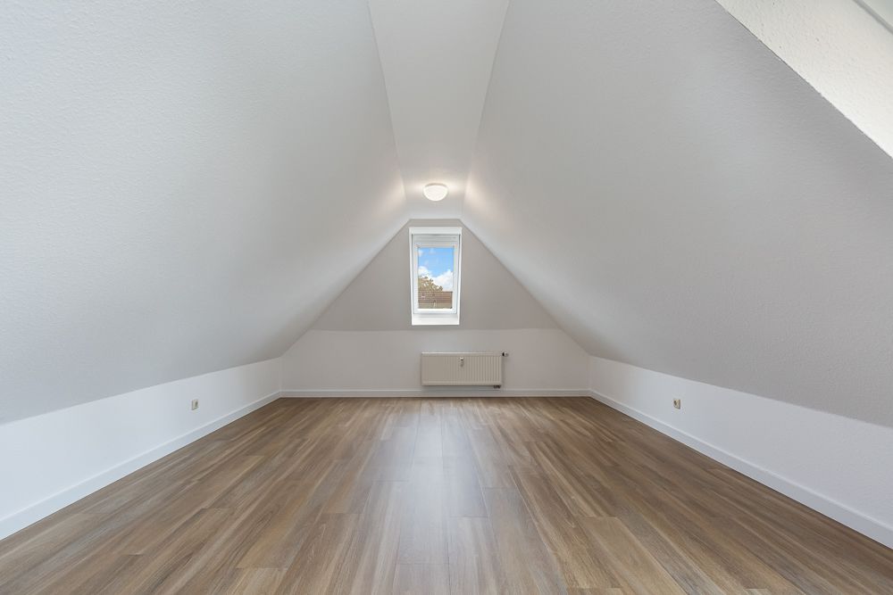 Leerer Dachgeschossraum mit schrägen weißen Wänden, Holzfußboden und einem Fenster mittig.