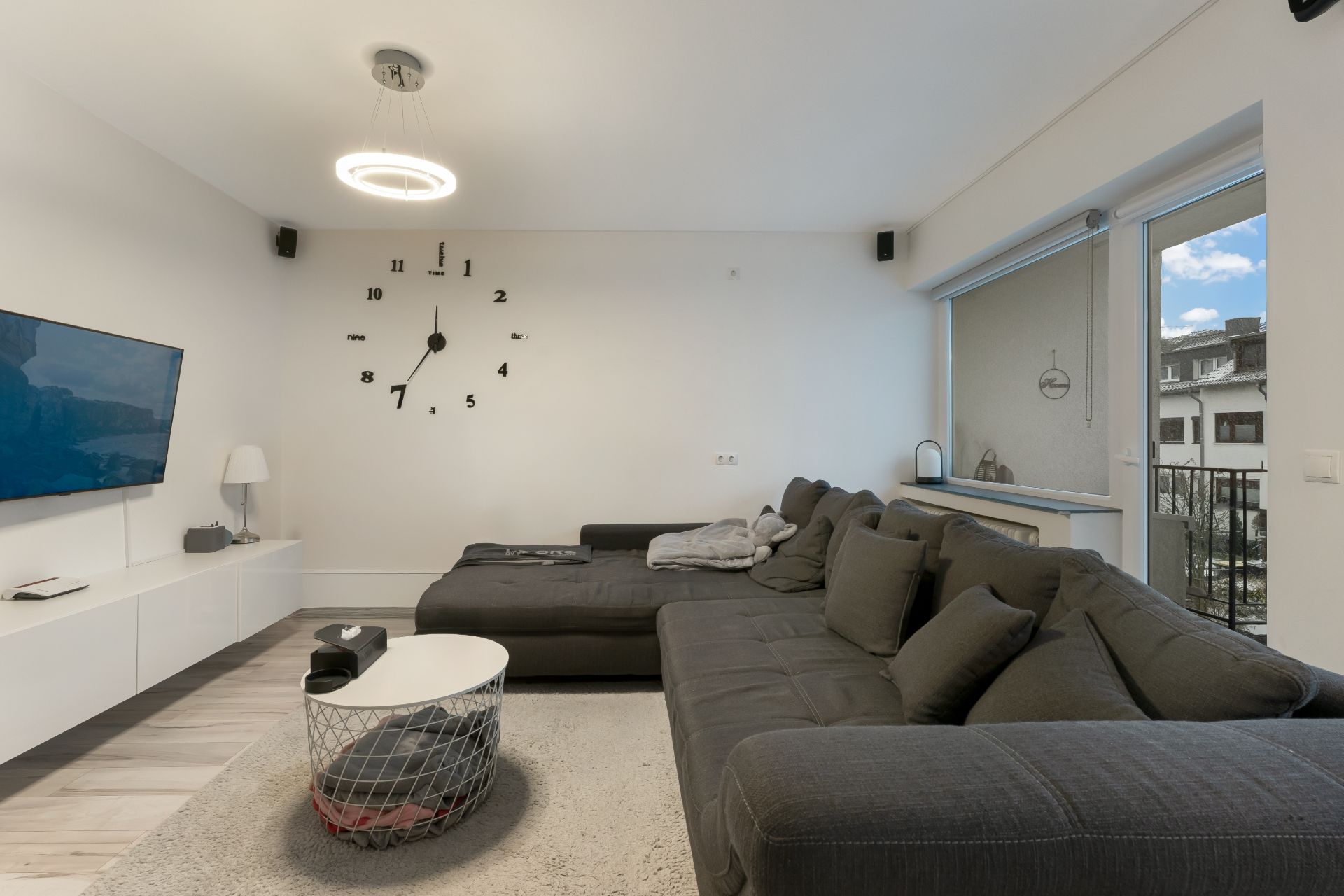Modernes Wohnzimmer mit grauem Sofa, Wand-Uhr, TV und großem Fenster.