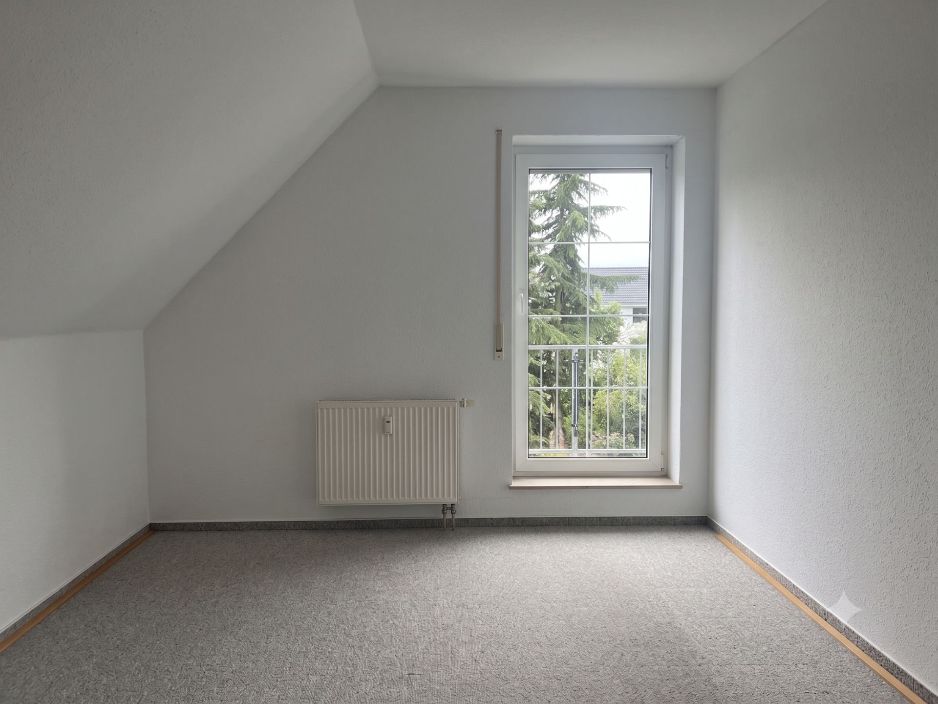Leeres Zimmer mit grauem Teppich, weißer Wand, Heizung und bodentiefem Fenster mit Blick ins Grüne.