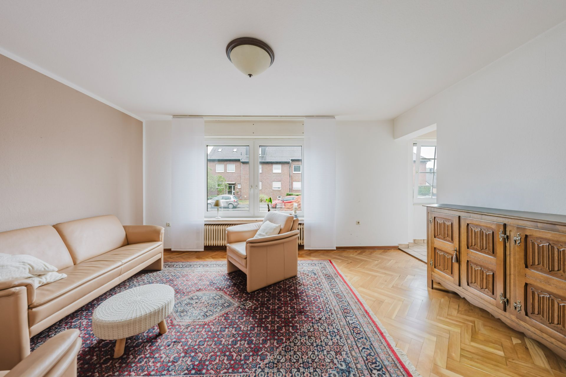 Wohnzimmer mit Teppich, Sofa, Sessel, Holzanrichte und großen Fenstern.