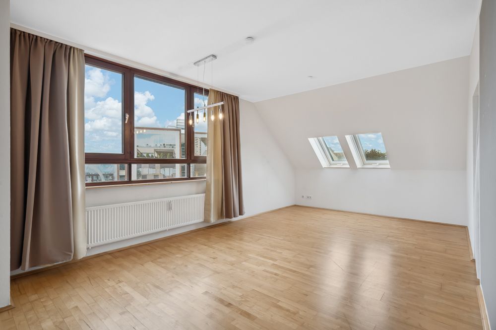 Helles, leeres Zimmer mit großen Fenstern, Parkettboden und Vorhängen.