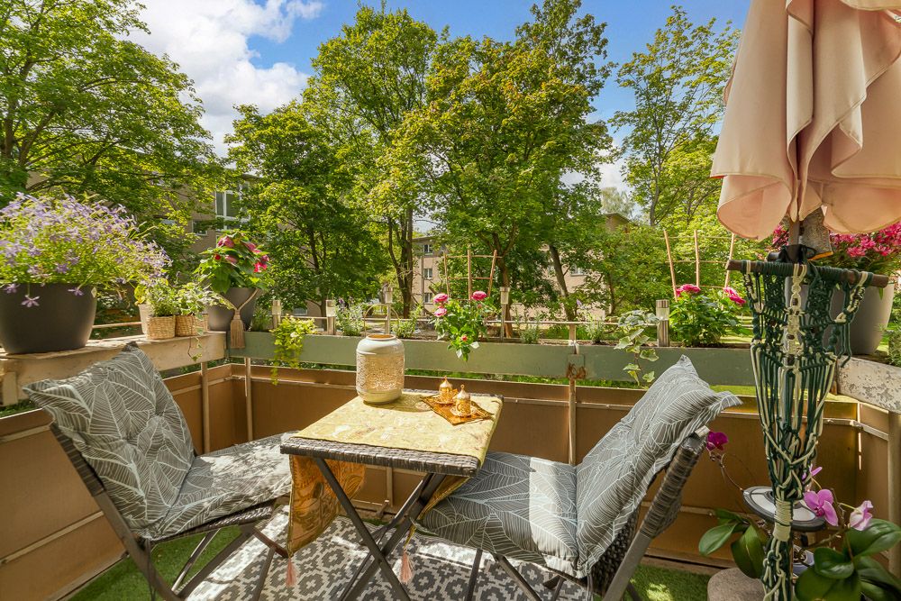 Balkon mit zwei Stühlen, Tisch, Pflanzen und Sonnenschirm, Blick auf grüne Bäume.
