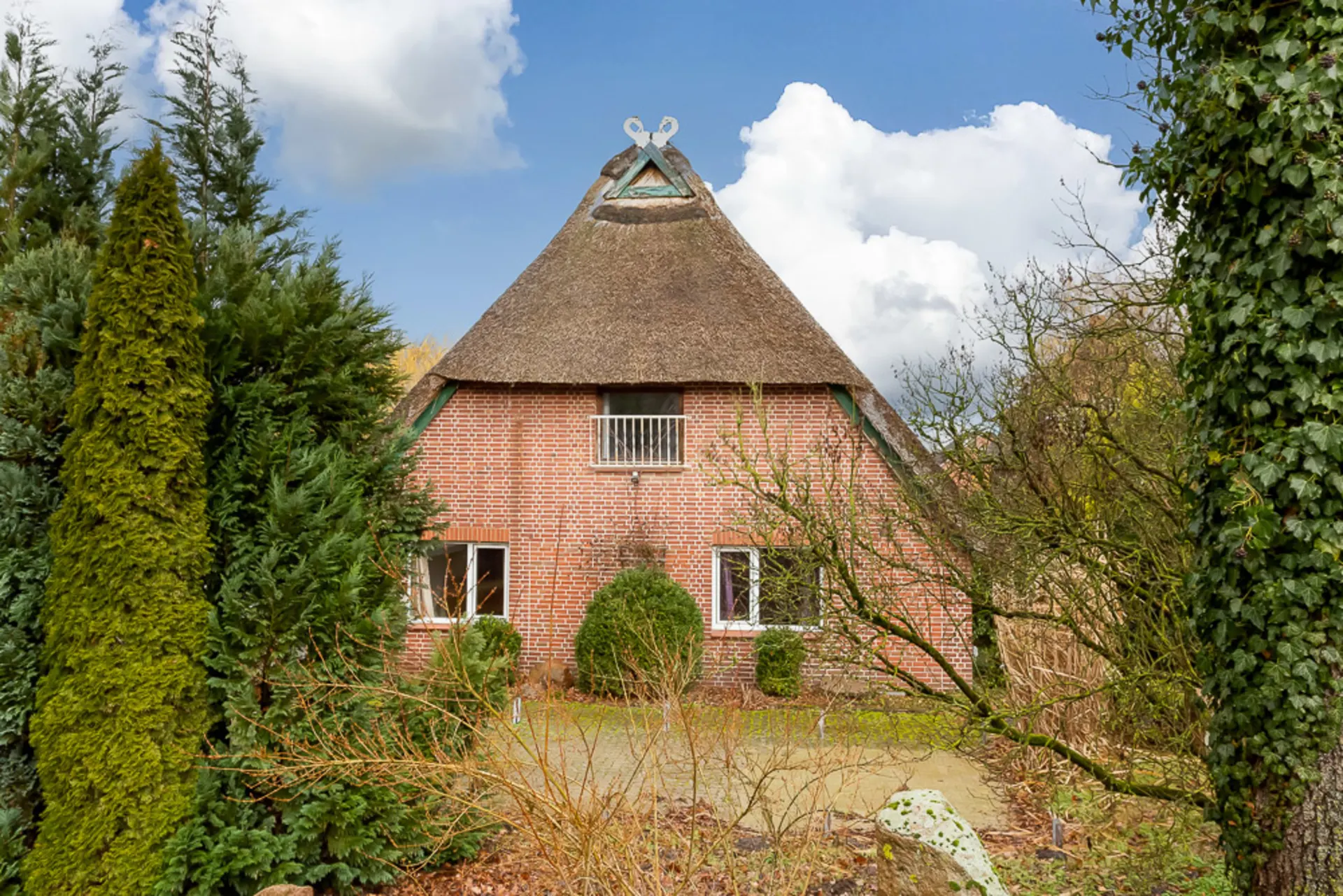 Backsteinhaus mit Reetdach und Garten, umgeben von Bäumen und Büschen.
