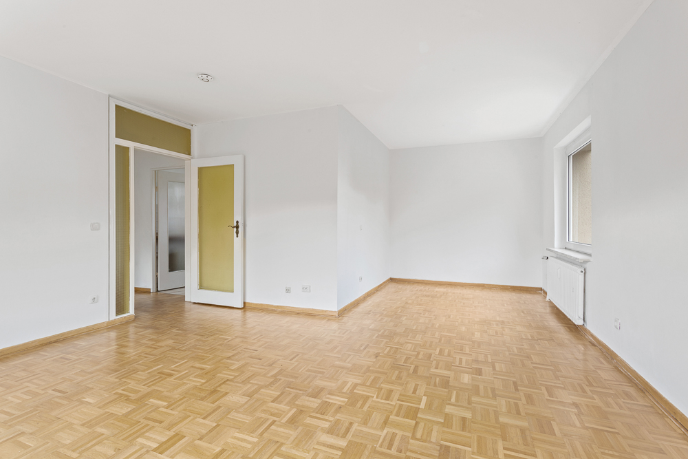 Leerer, heller Raum mit Parkettboden, weißer Wand, offenem Fenster und offener Tür.