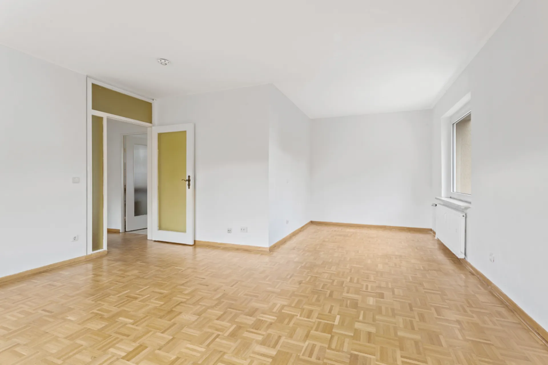 Leerer, heller Raum mit Parkettboden, weißer Wand, offenem Fenster und offener Tür.