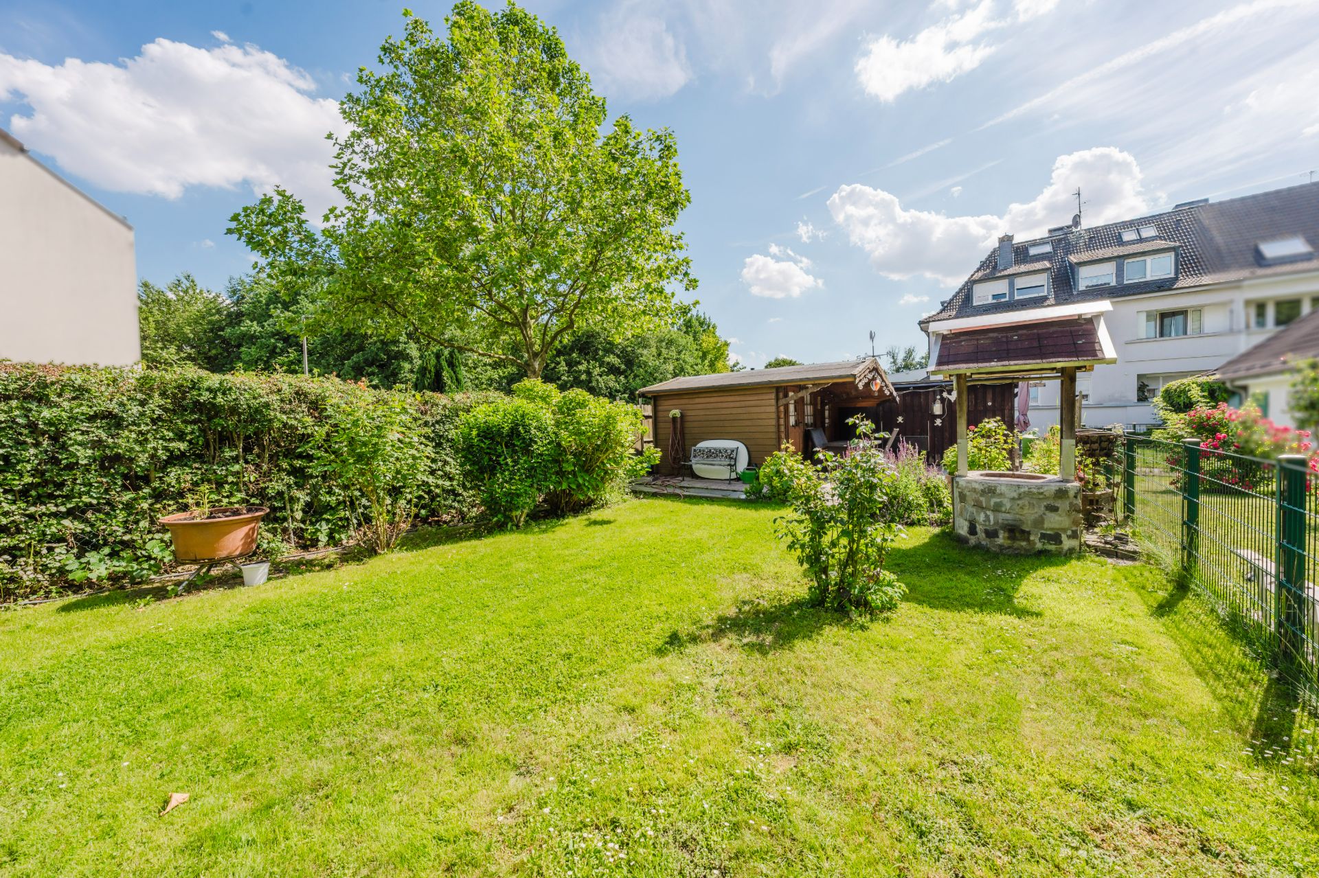 Grüner Garten mit Rasen, Sträuchern, Brunnen und kleinem Gartenhaus, daneben ein Wohnhaus.
