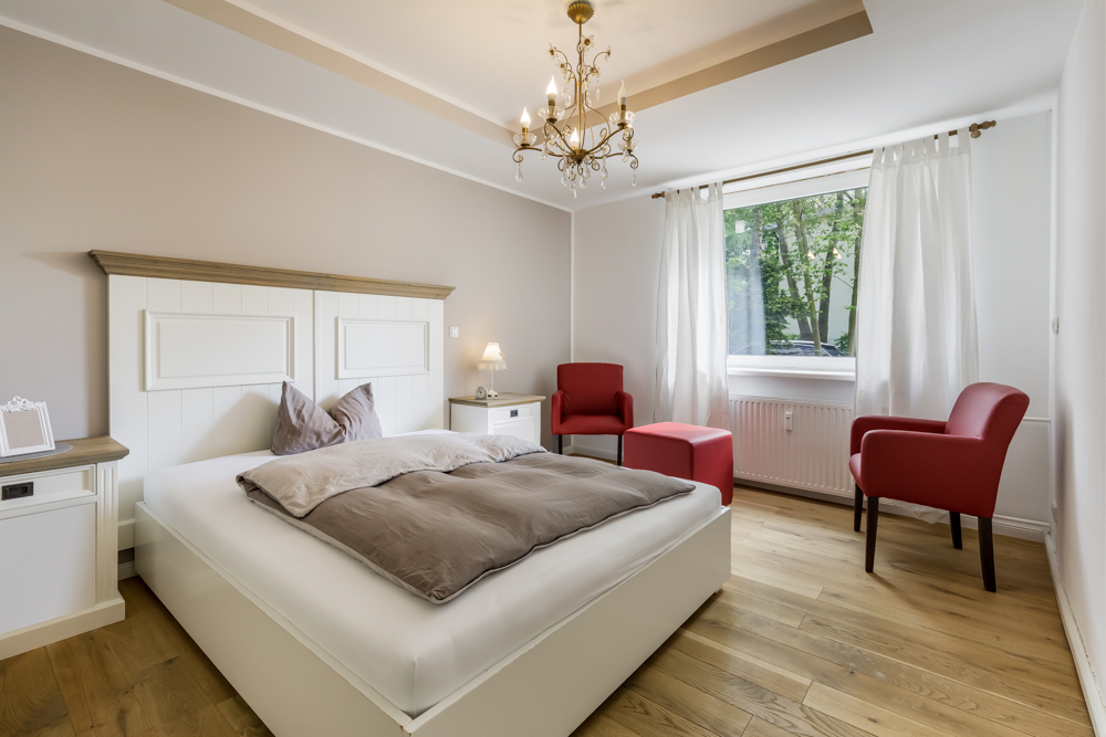 Helles Schlafzimmer mit Doppelbett, zwei roten Sesseln, Holzboden und großem Fenster.