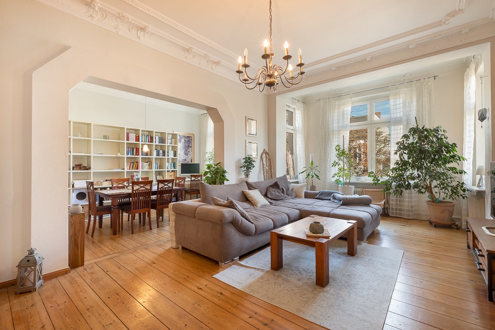 Helles Wohnzimmer mit großem Ecksofa, Holztisch, Essbereich, Bücherregal und vielen Pflanzen.