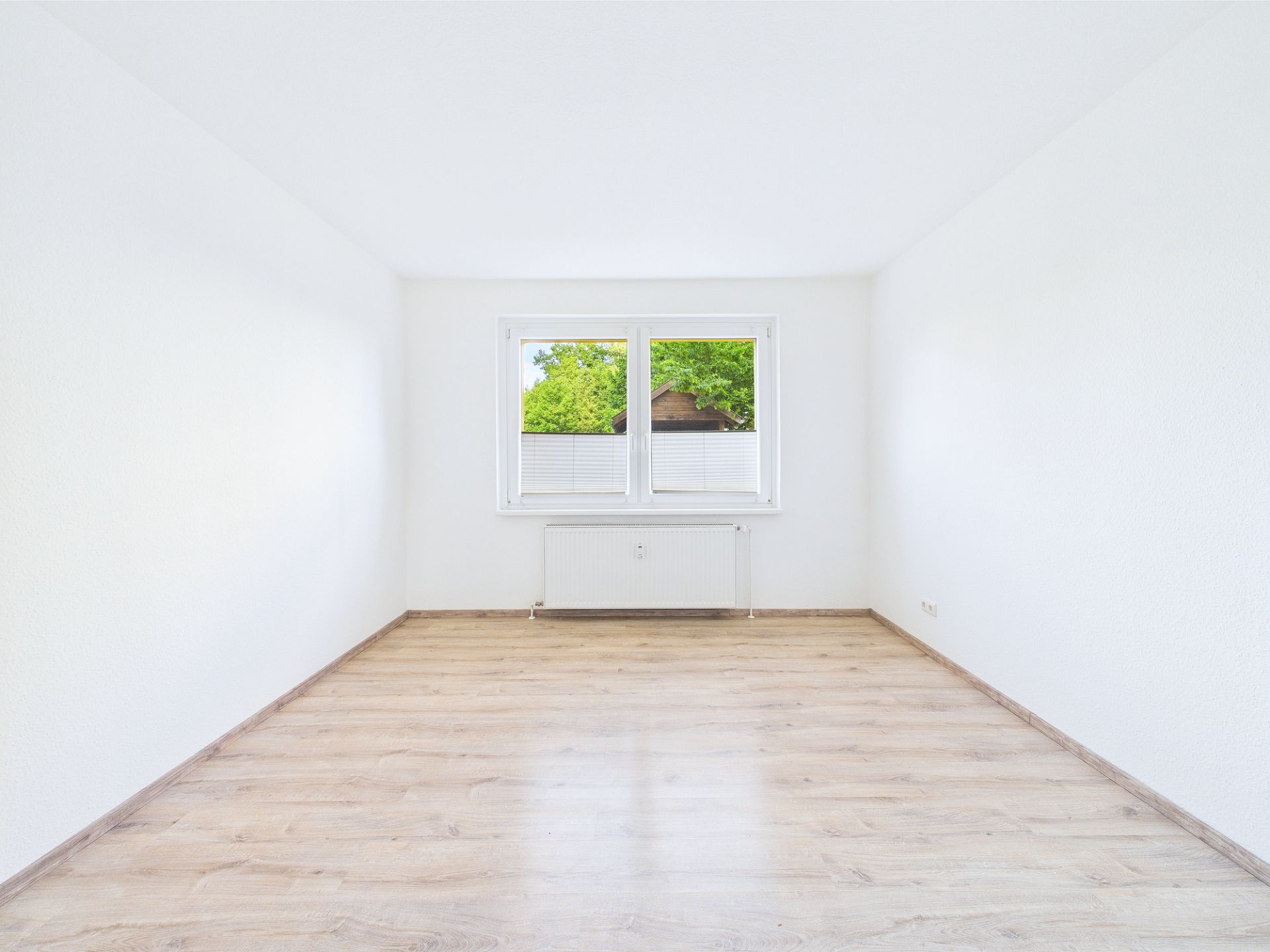 Leerer, heller Raum mit Holzfußboden und zwei Fenstern, durch die Bäume draußen sichtbar sind.