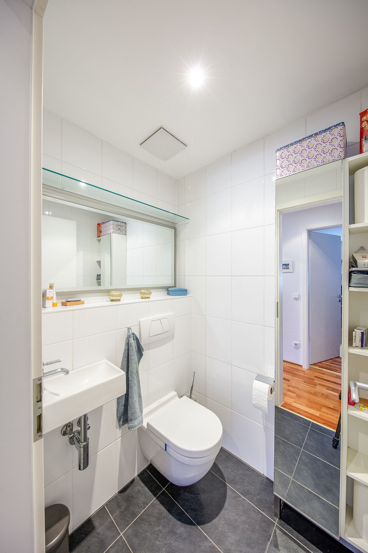 Modernes Badezimmer mit Wandtoilette, kleinem Waschbecken und Spiegel, helle Fliesen.