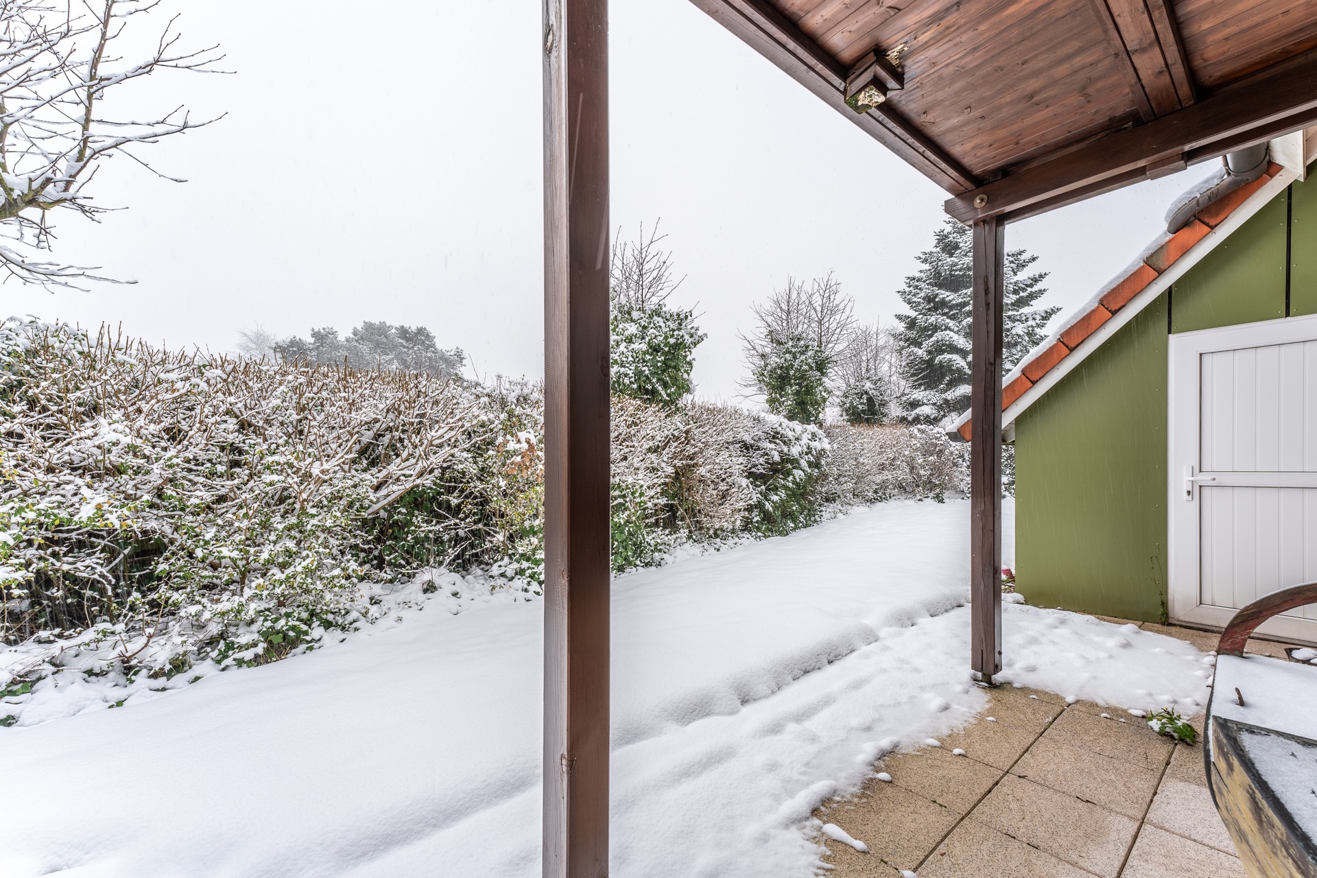 Schneebedeckter Garten mit Terrasse und grünem Haus bei winterlichem Wetter.