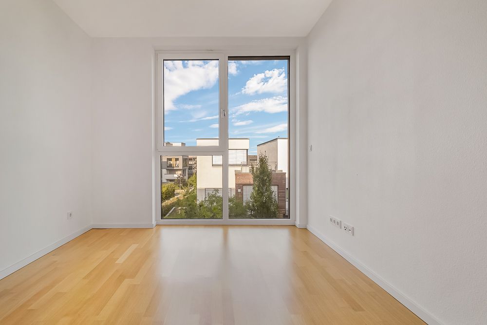 Leerer Raum mit hellem Holzboden und großem Fenster, Blick auf moderne Wohnhäuser.