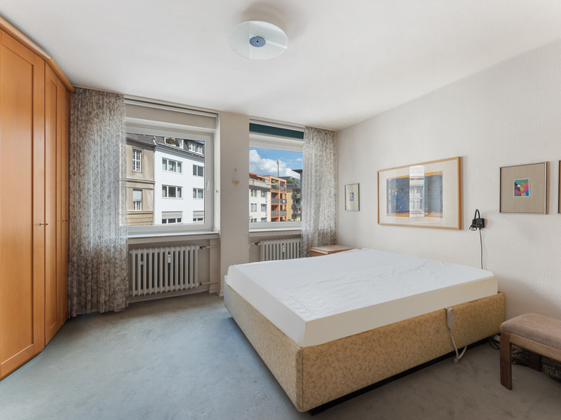 Helles Schlafzimmer mit großem Bett, Holzschrank und zwei Fenstern mit Blick auf andere Gebäude.
