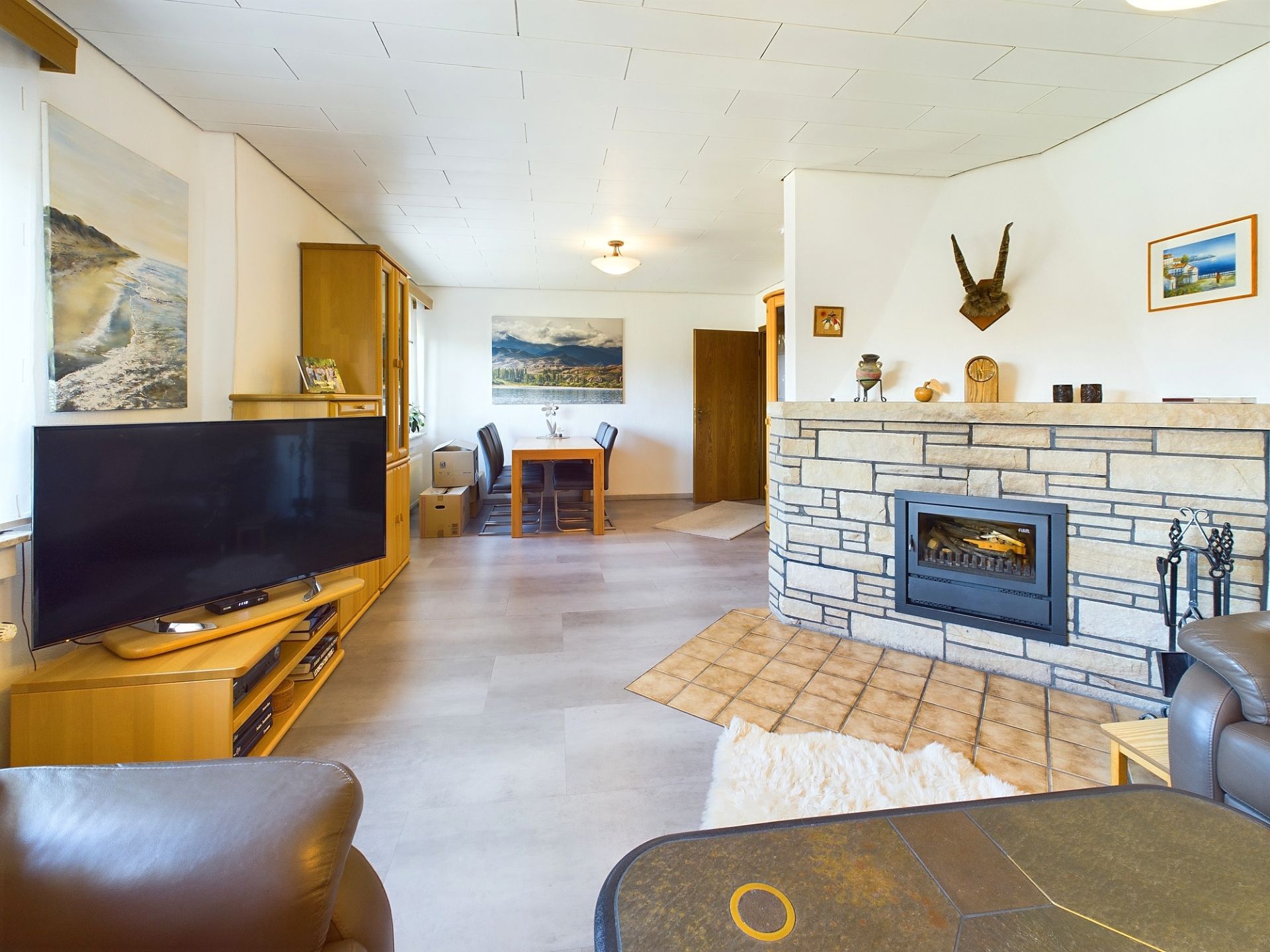 Wohnzimmer mit Fernseher, Kamin und Essbereich, helle Einrichtung und dekorative Bilder an den Wänden.