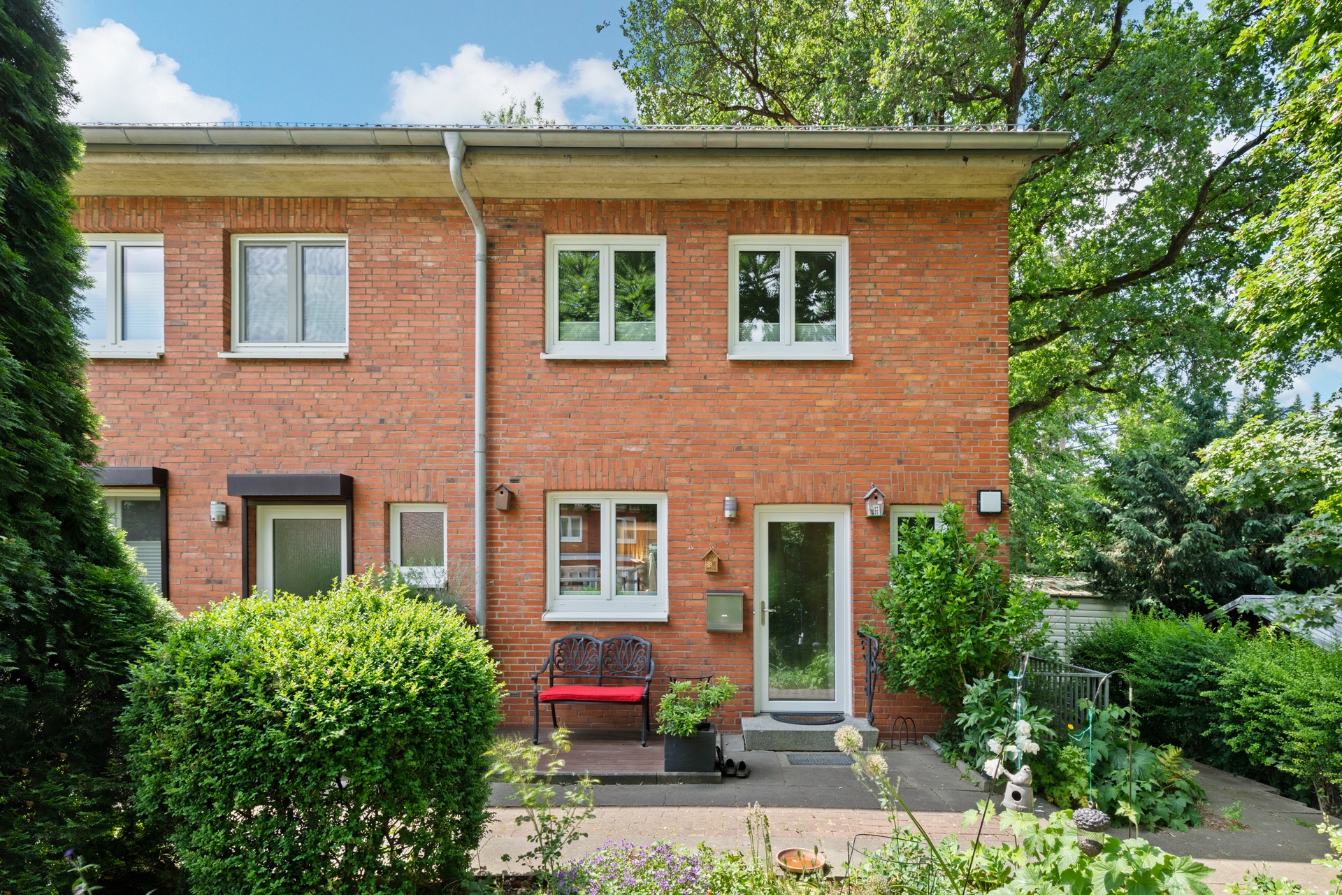 Rotes Backsteinhaus mit weiß gerahmten Fenstern, kleiner Garten und Bank vor dem Eingang.