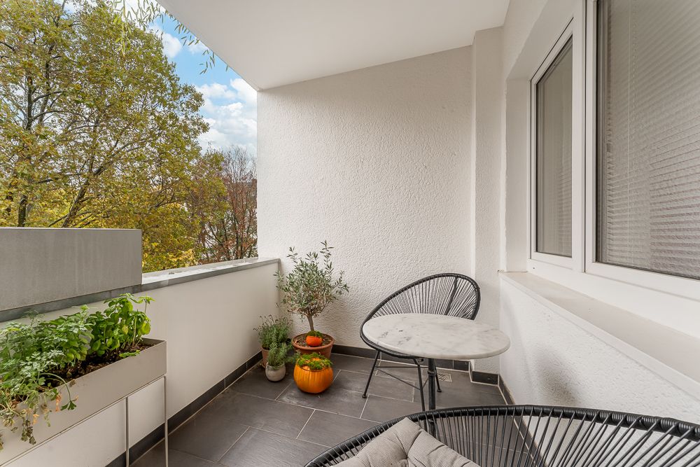 Balkon mit zwei Stühlen, rundem Tisch, Pflanzkasten und Topfpflanzen, Blick auf Bäume.