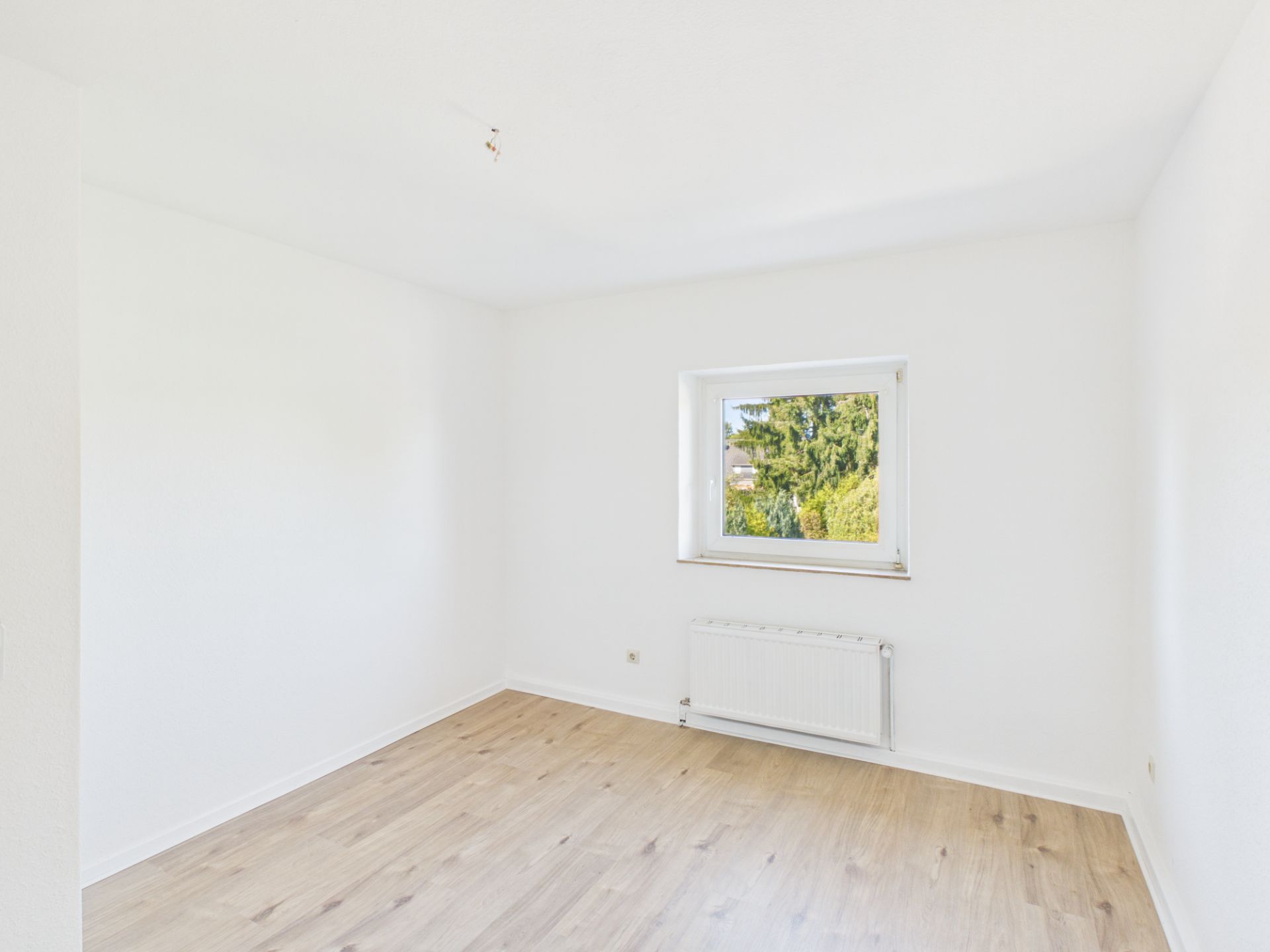 Leerer, heller Raum mit Holzboden, weißer Wand, Fenster und Heizkörper.
