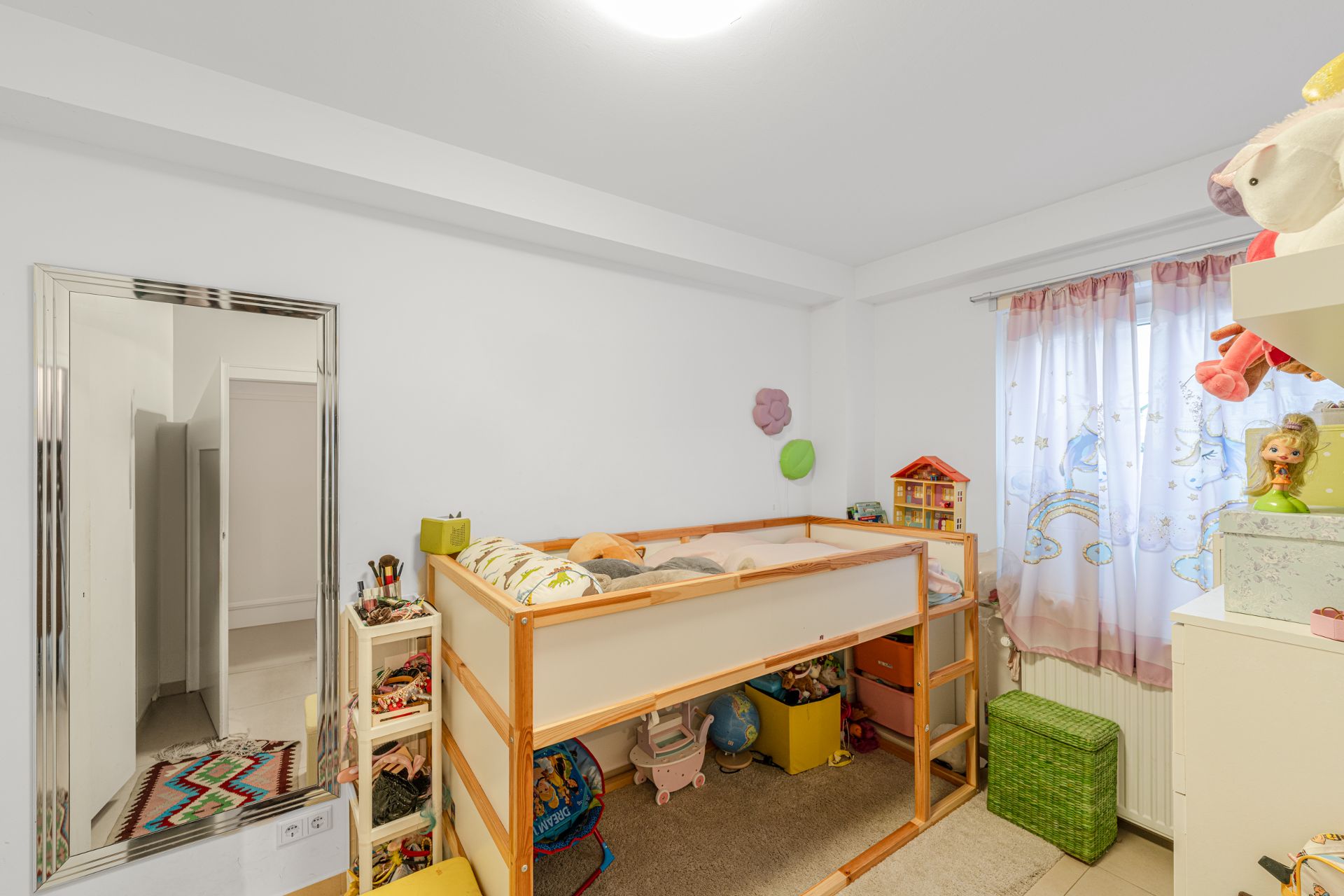 Kinderzimmer mit Hochbett, Spielzeugregalen und bunten Vorhängen vor dem Fenster.