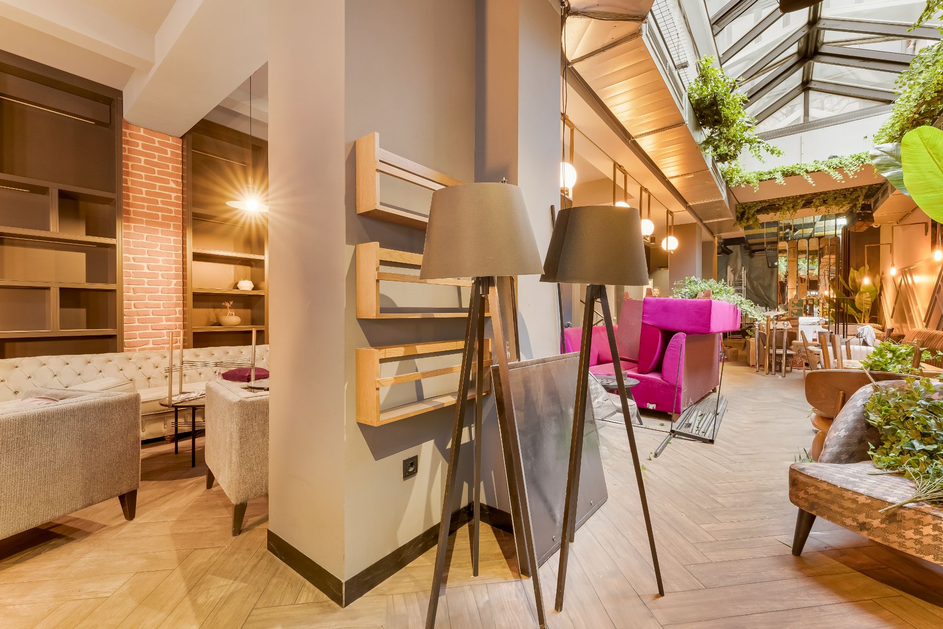 Wohnzimmer mit Stehlampen, grauen Sofas, pinkem Sessel und viel Pflanzen unter Glasdach.