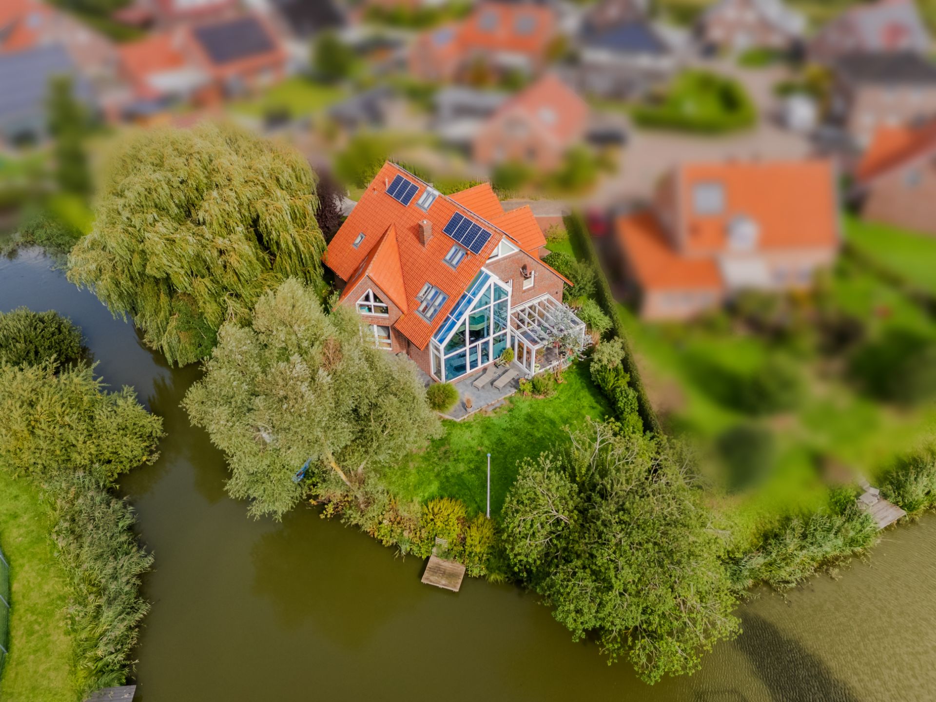 Luftaufnahme eines Hauses mit roten Dächern am Wasser, umgeben von Bäumen und Garten.