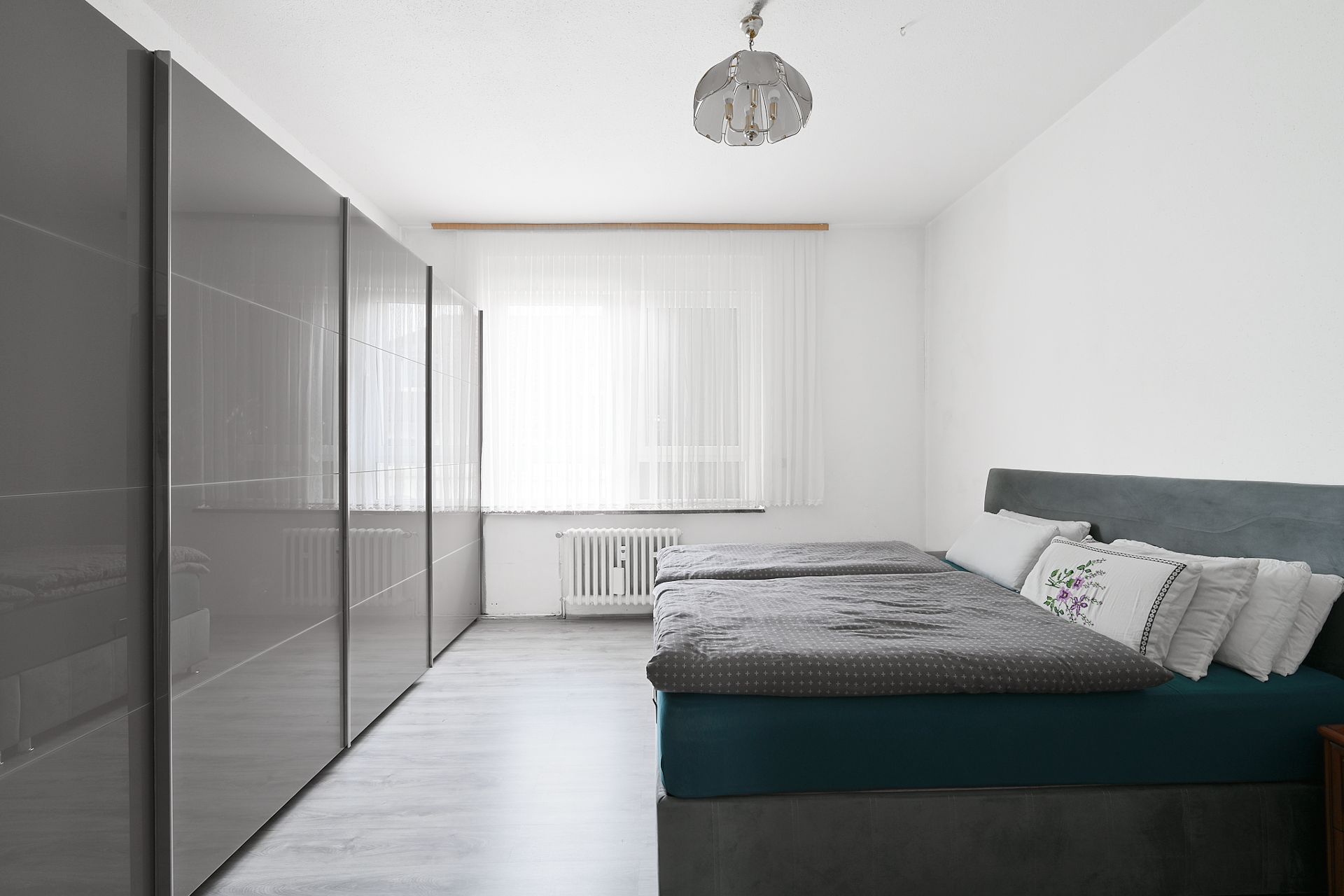 Helles Schlafzimmer mit großem Bett, grauem Kleiderschrank und Fenster mit weißen Vorhängen.