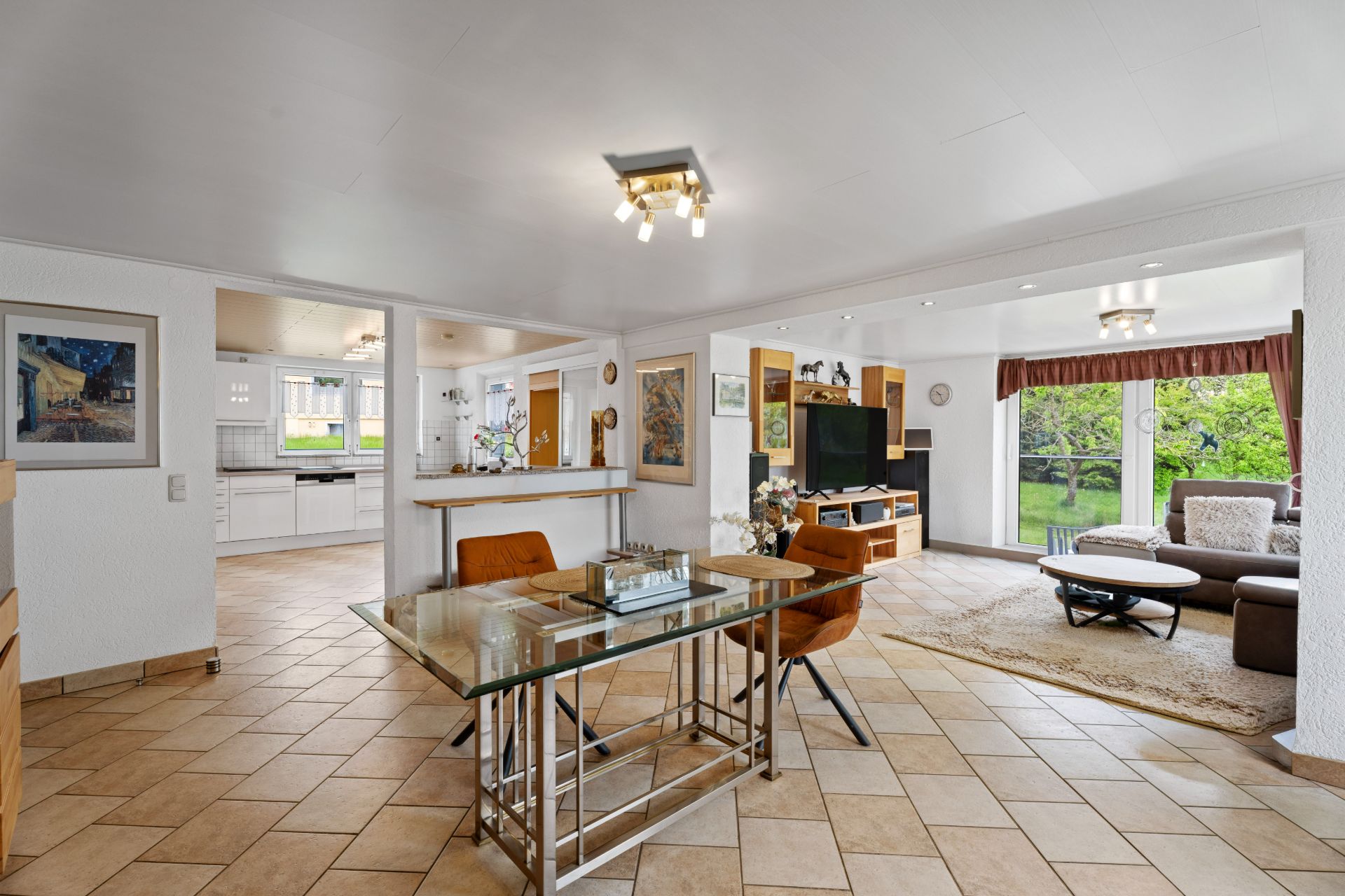Helles Wohnzimmer mit Glastisch, braunen Stühlen, Sofa, Fernseher und Blick auf Garten.