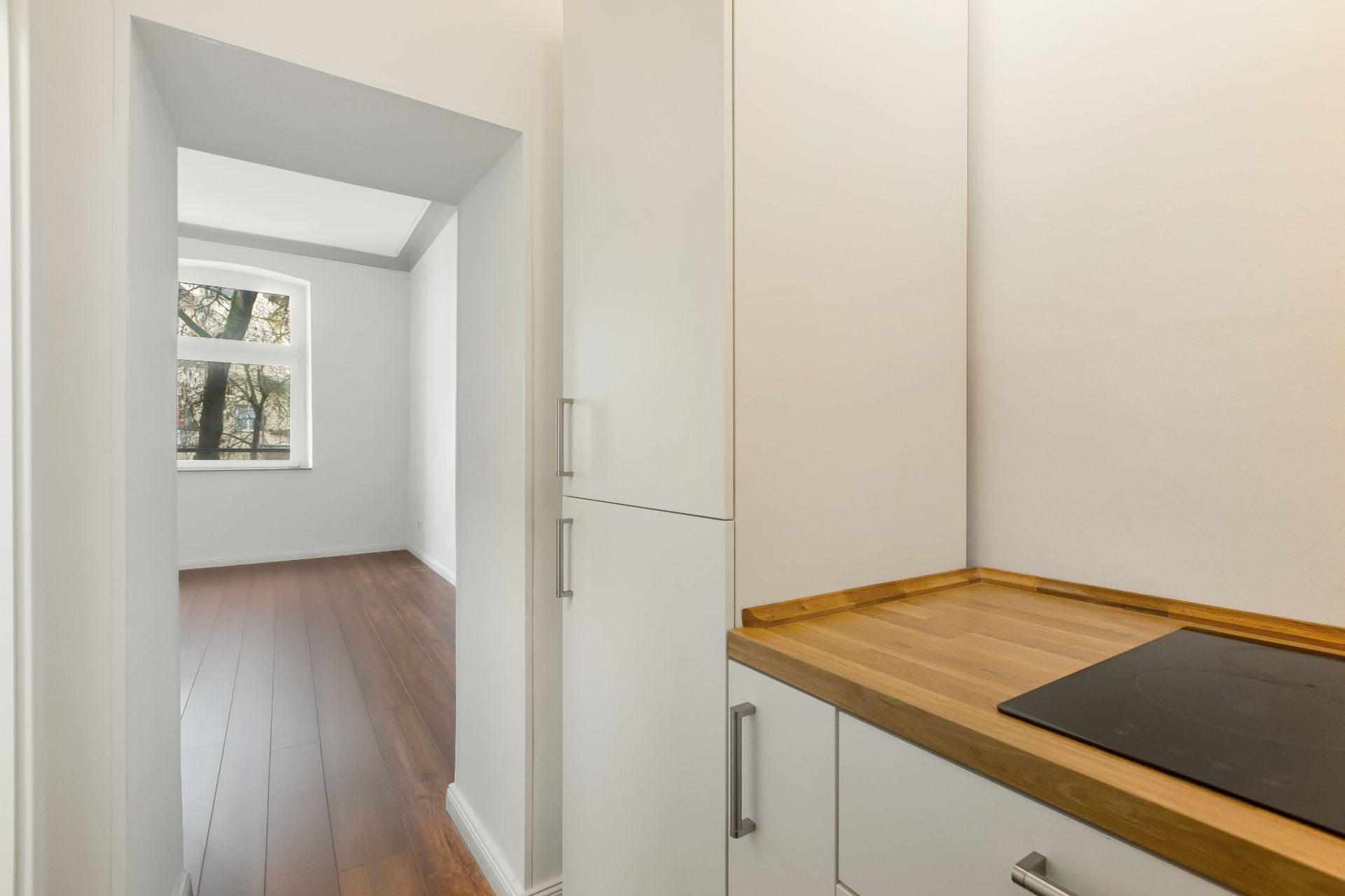Helle Küche mit Holzarbeitsplatte, daneben ein Raum mit dunklem Holzboden und Fenster.