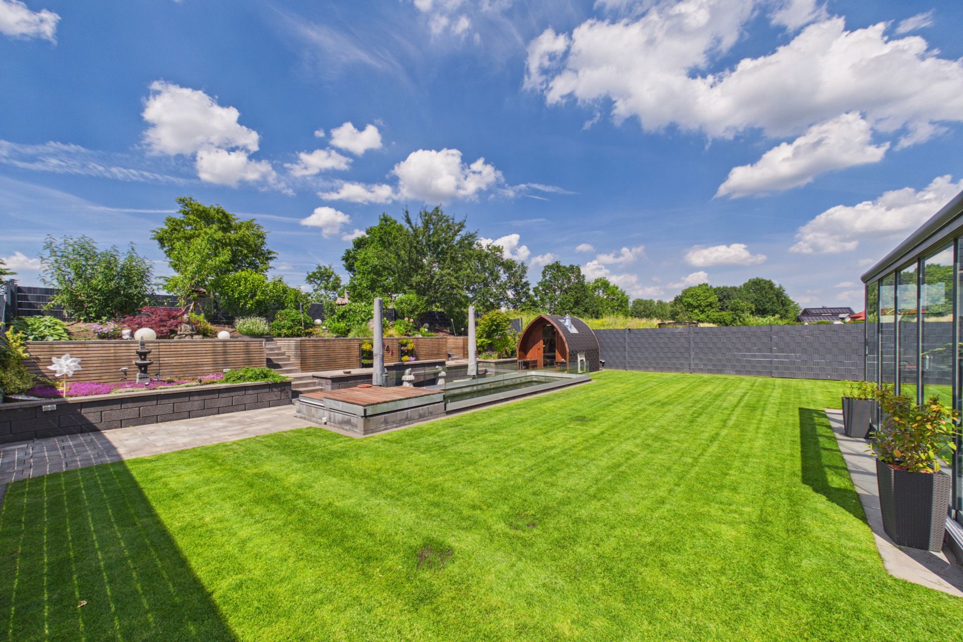 Großer Garten mit Rasen, Terrasse, Teich und moderner Begrenzungsmauer.