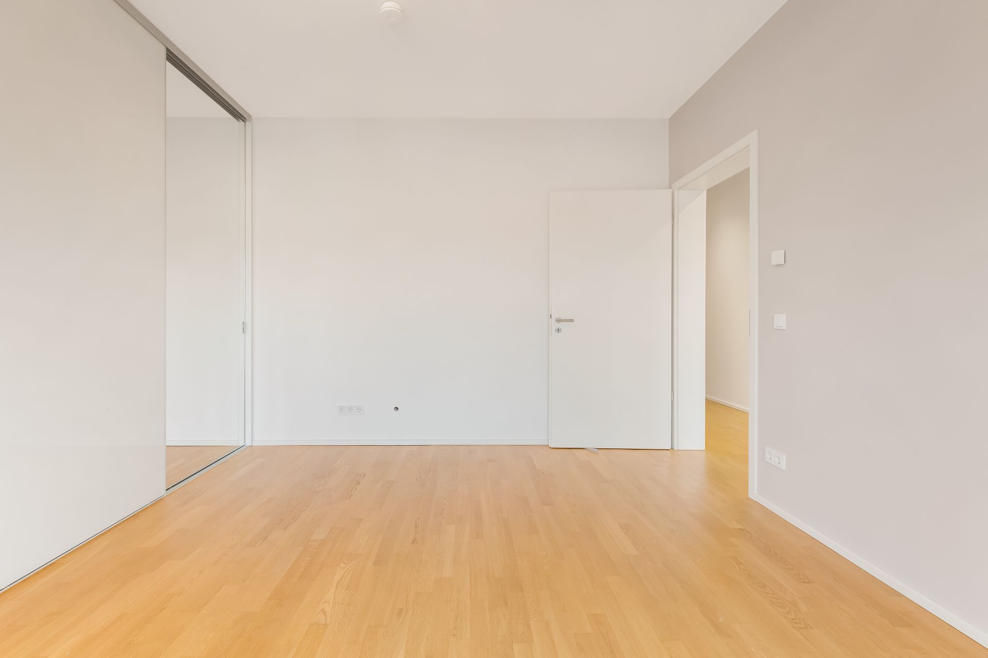 Leerer Raum mit hellem Holzboden, weißen Wänden und offener Tür, Spiegel an der linken Wand.