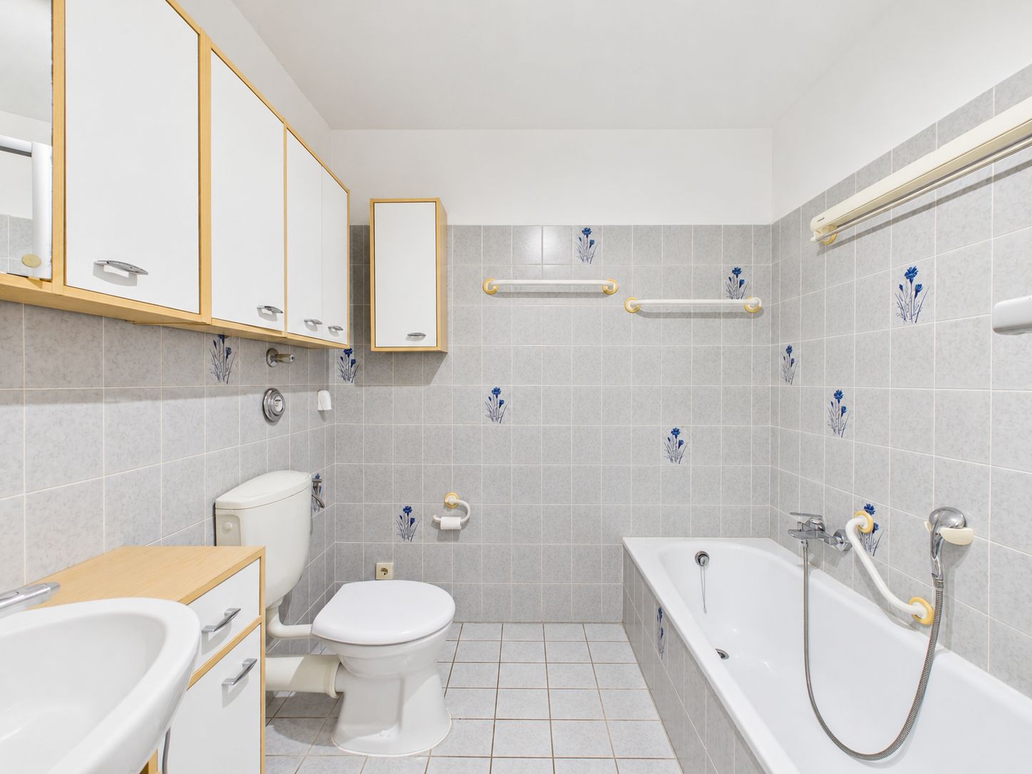 Helles Badezimmer mit Badewanne, Toilette, Waschbecken und grauen Fliesen mit Blumenmuster.