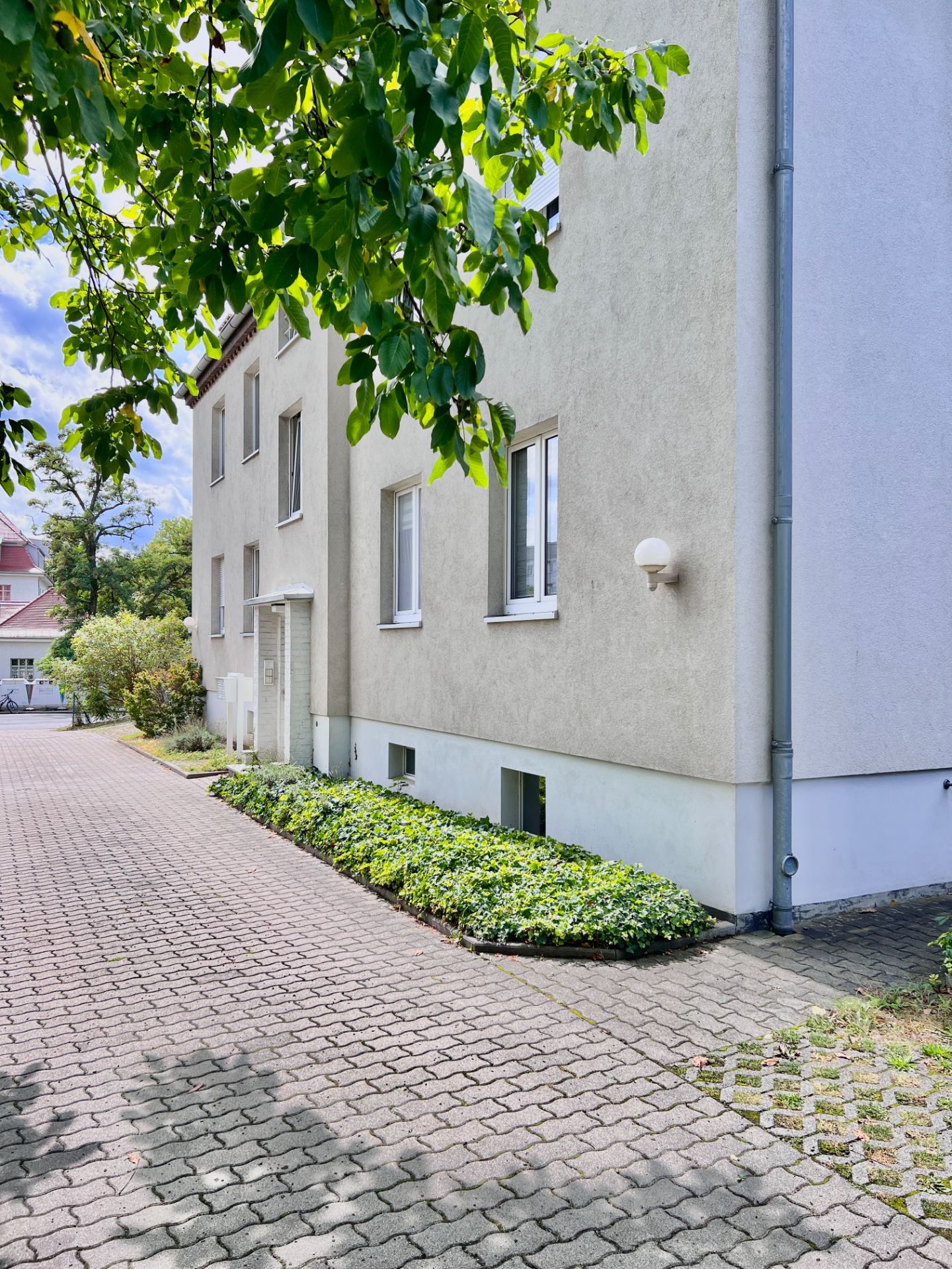 Mehrfamilienhaus mit Pflasterweg, Beeten und großen grünen Bäumen im Vordergrund.
