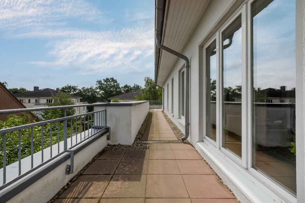 Lange Terrasse mit Fliesenboden und Geländer an einem Wohngebäude, große Fenster auf der rechten Seite.