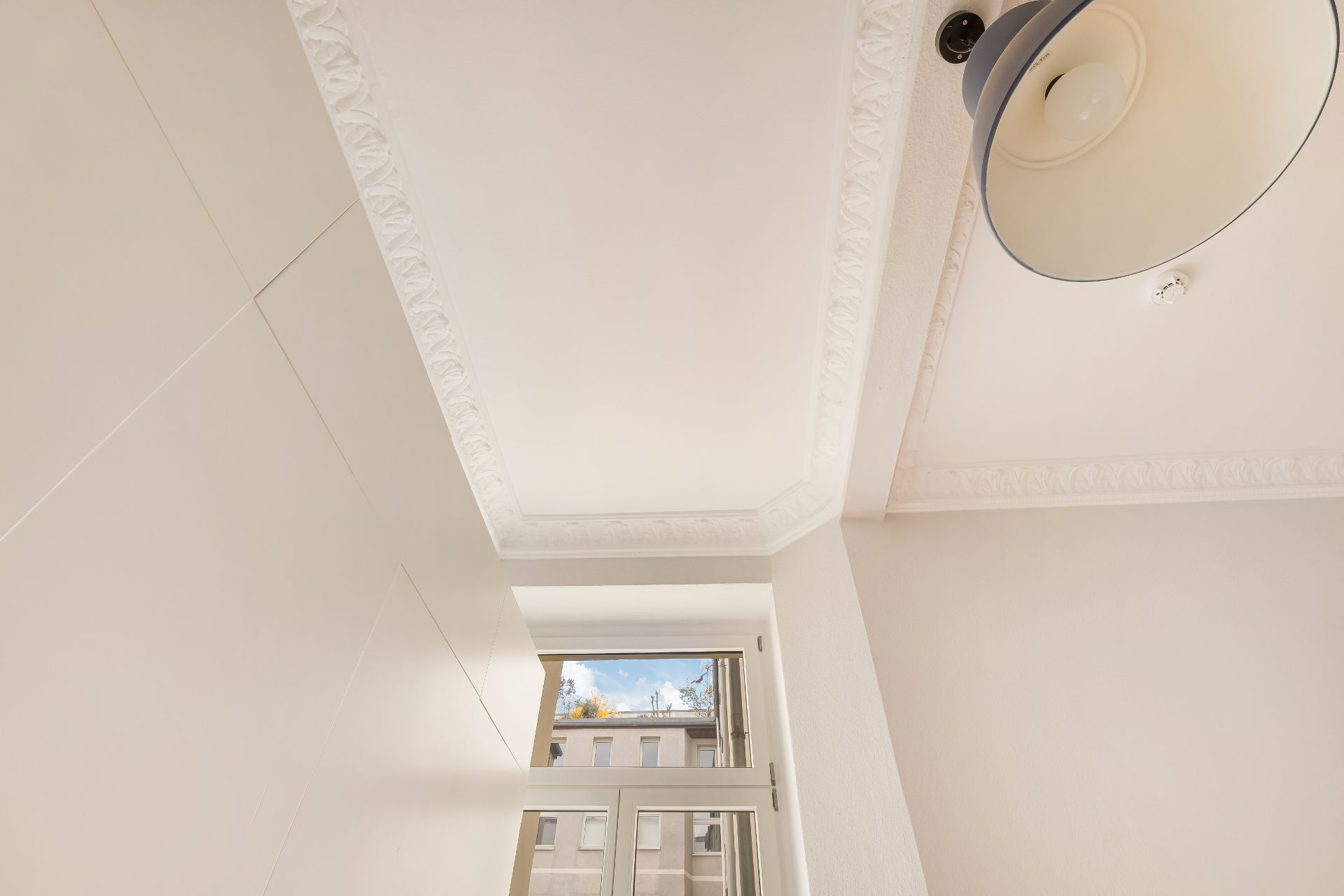 Weiße Zimmerdecke mit Stuckverzierung und Deckenlampe, darunter ein Fenster.