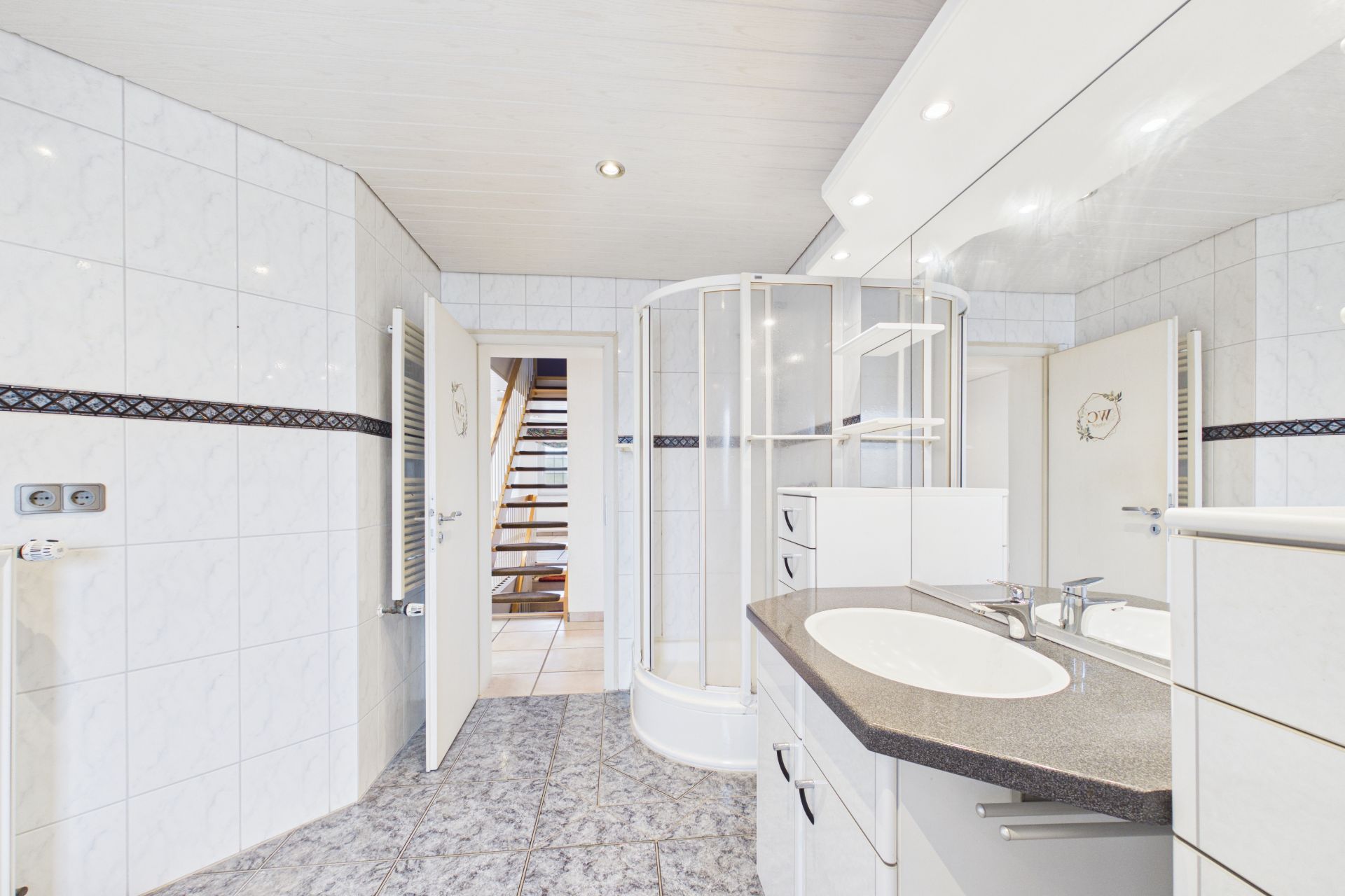 Helles Badezimmer mit Dusche, großem Spiegel und Waschbecken, sowie Blick auf eine offene Tür.