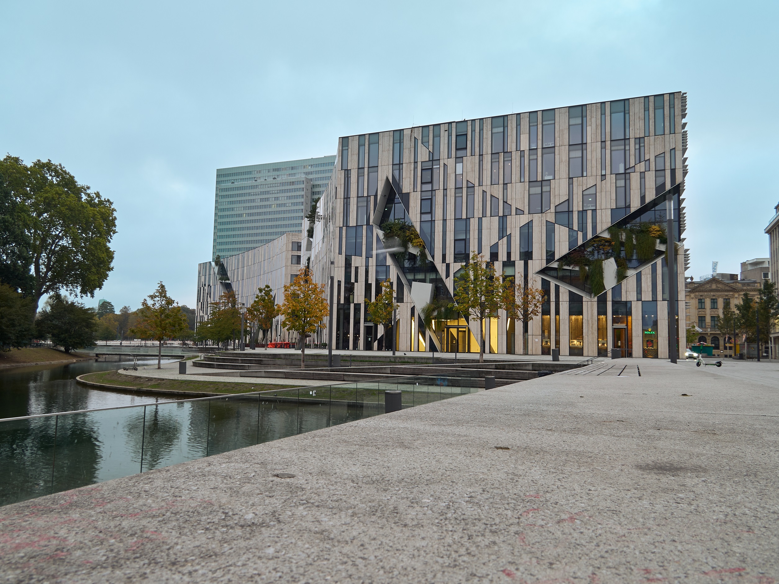 Modernes Gebäude mit großen Fenstern und geometrischen Mustern neben einem Wasserbecken und Bäumen.