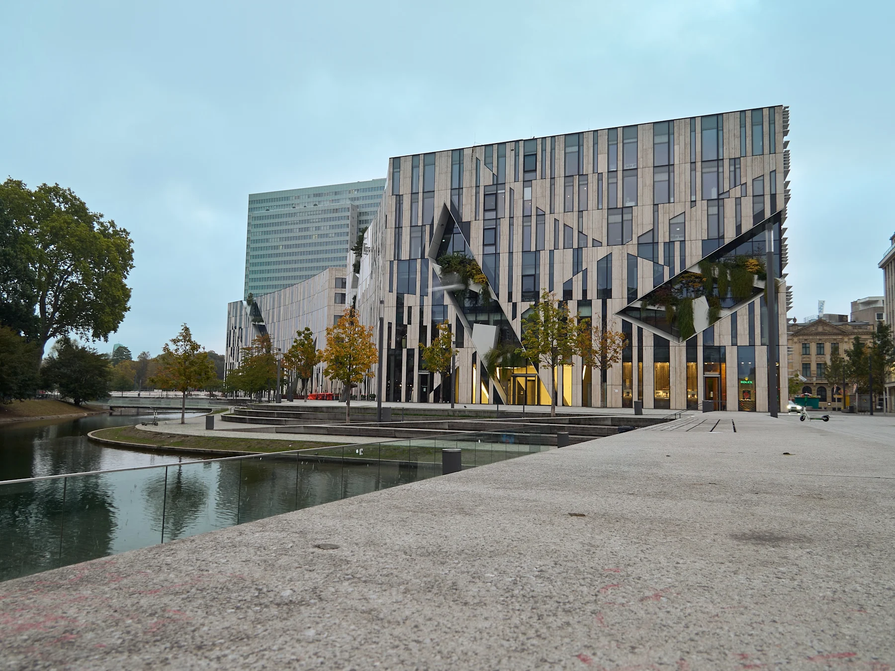 Modernes Gebäude mit großen Fenstern und geometrischen Mustern neben einem Wasserbecken und Bäumen.