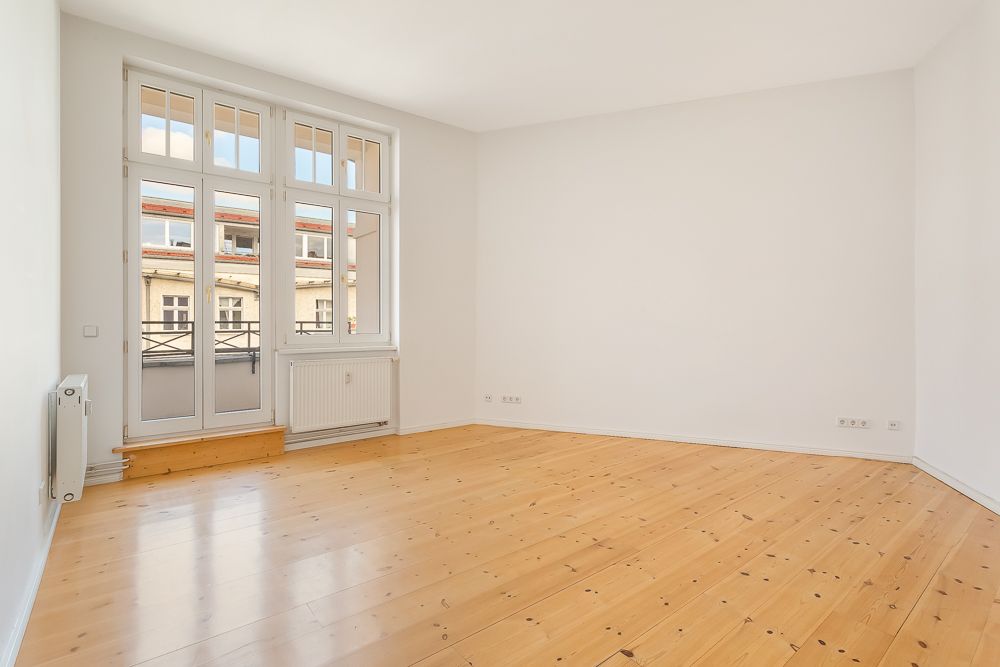 Leerer Raum mit hellem Holzboden, weißen Wänden und großen Fenstern mit Zugang zum Balkon.