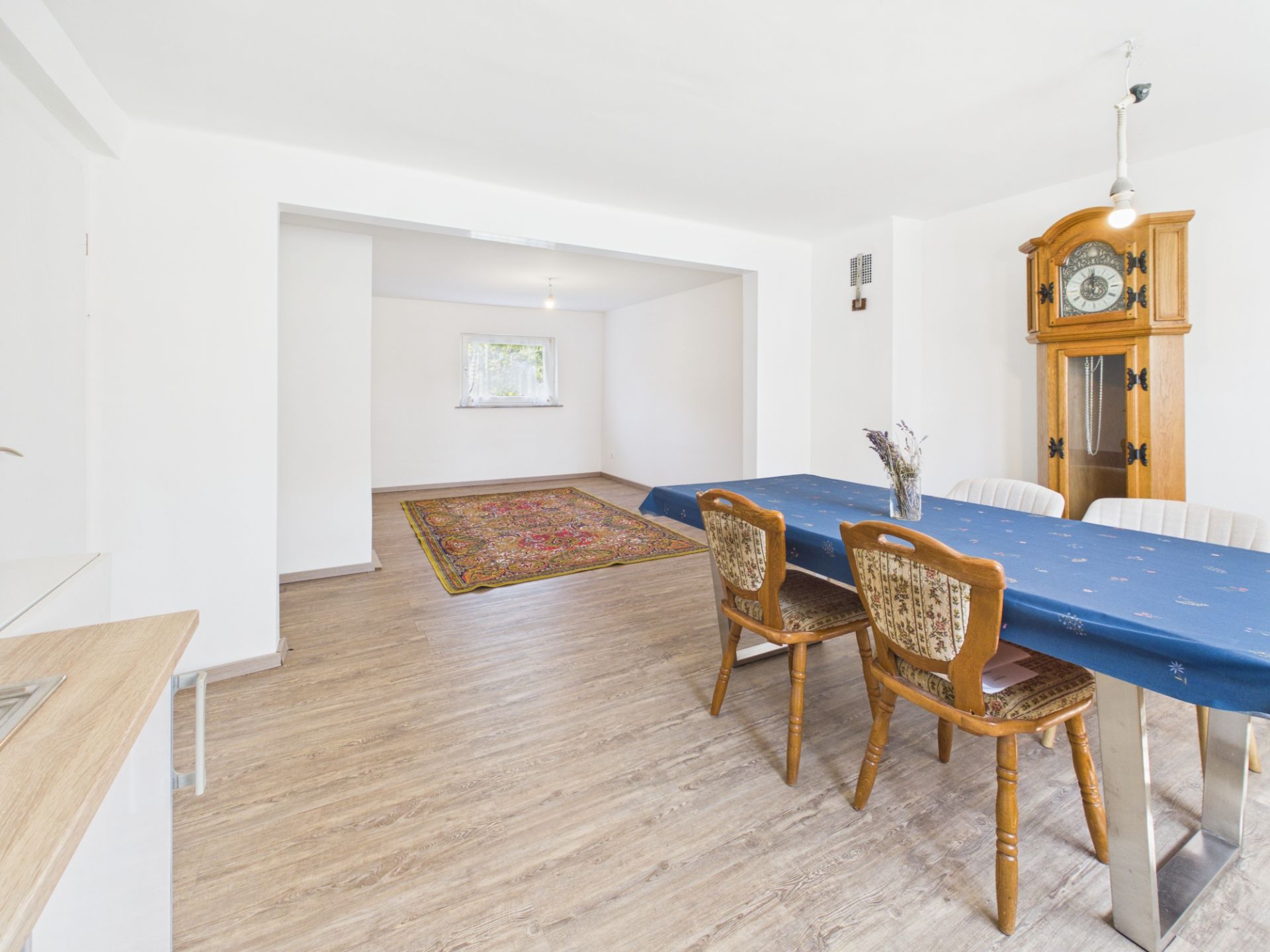 Esszimmer mit blauem Tisch, vier Stühlen, Standuhr und angrenzendem Raum mit Teppich.
