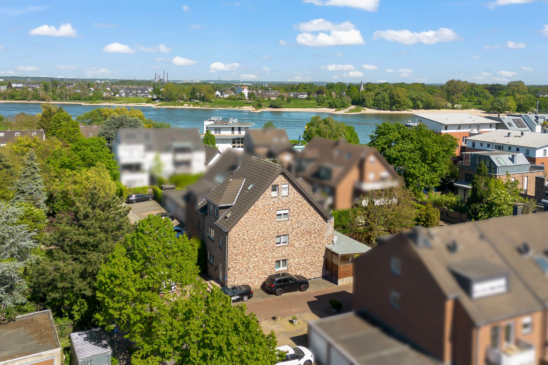 Backsteinwohnhaus mit Garten, umgeben von Bäumen, im Vordergrund Autos, Fluss im Hintergrund.