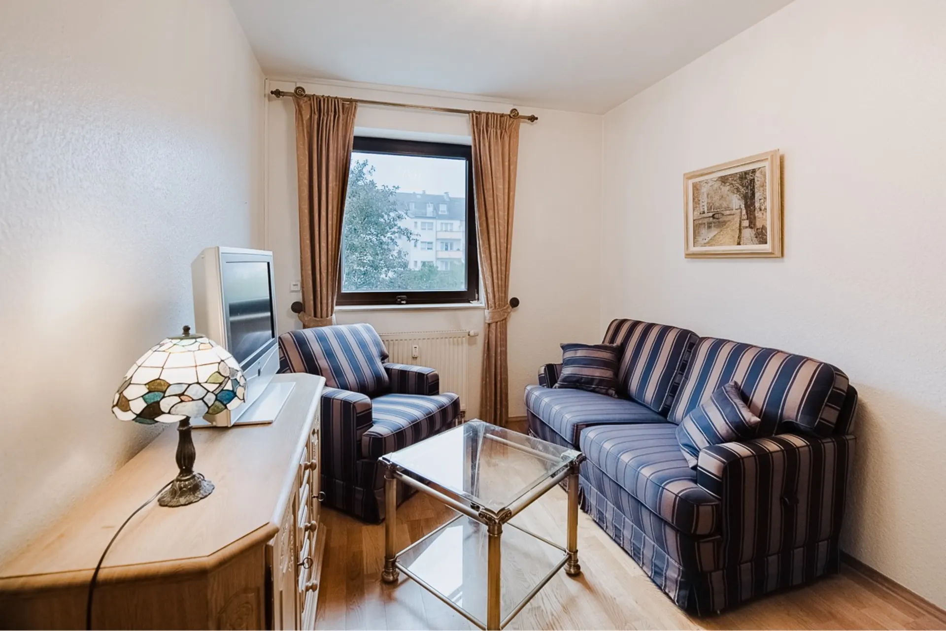 Kleines Wohnzimmer mit gestreiftem Sofa, Sessel, Glastisch und Fernseher vor Fenster.